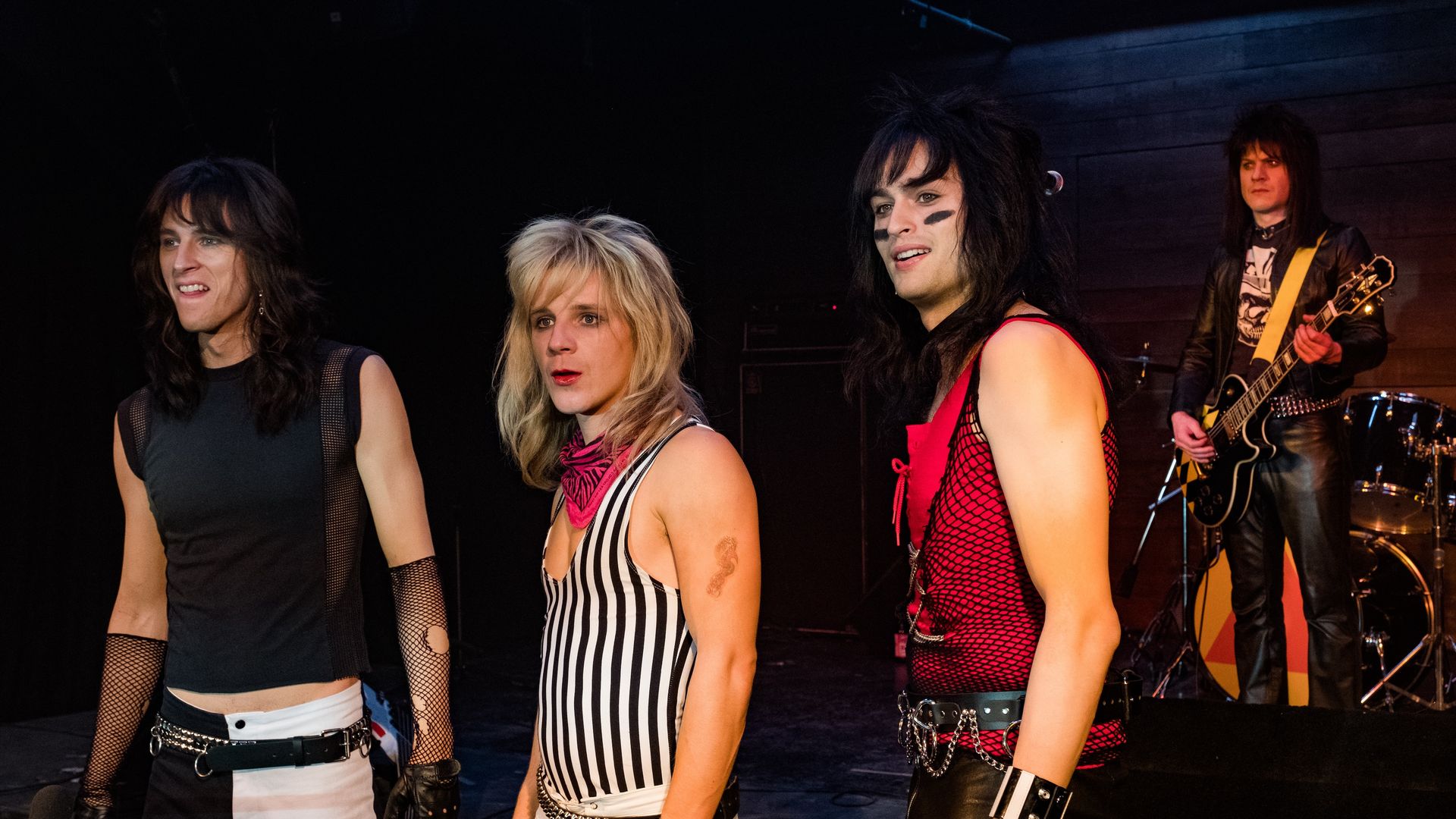 Four rock performers on a dark stage: left woman in black top; blonde singer in striped dress; guitarist with hair and face paint in red fishnet; far right guitarist in leather with yellow strap.