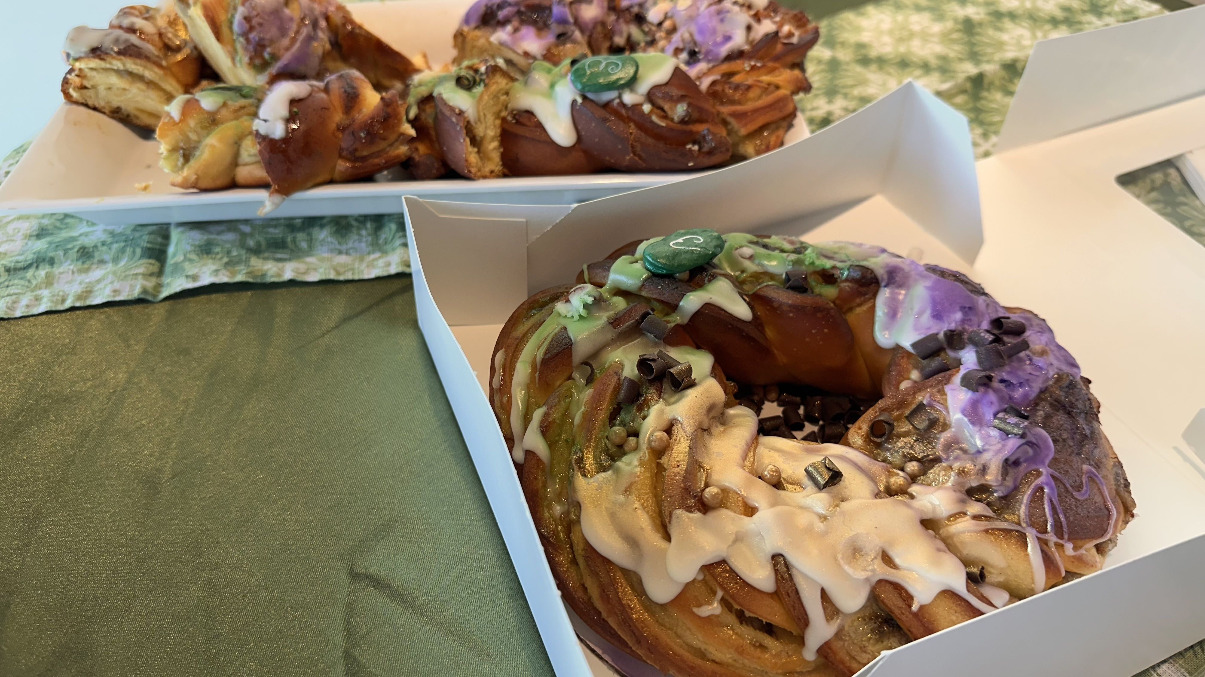 Two braided cakes on a green tablecloth, each topped with white glaze, , purple, green and gold sugar, chocolate chips, and small decorative pearls.