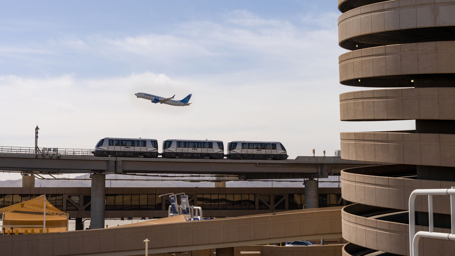 United Airlines plane taking off in clear sky above three connected white airport shuttle cars on elevated track near a circular beige parking structure.