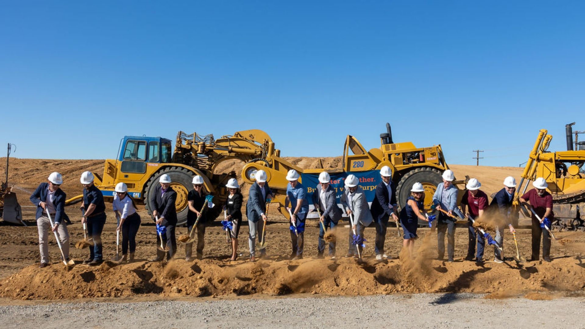 Group of people in hard hats and formal attire with shovels breaking ground at construction site with large yellow machinery under clear blue sky.