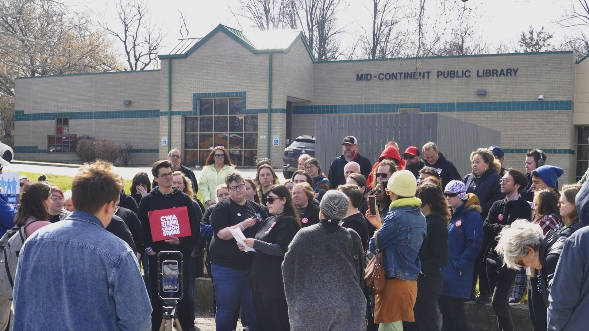 Crowd gathers outside a beige-brick building labeled Mid-Continent Public Library for a rally. Attendees wear jackets and hats; a red sign reads CWA STRONG UNION STRONG.