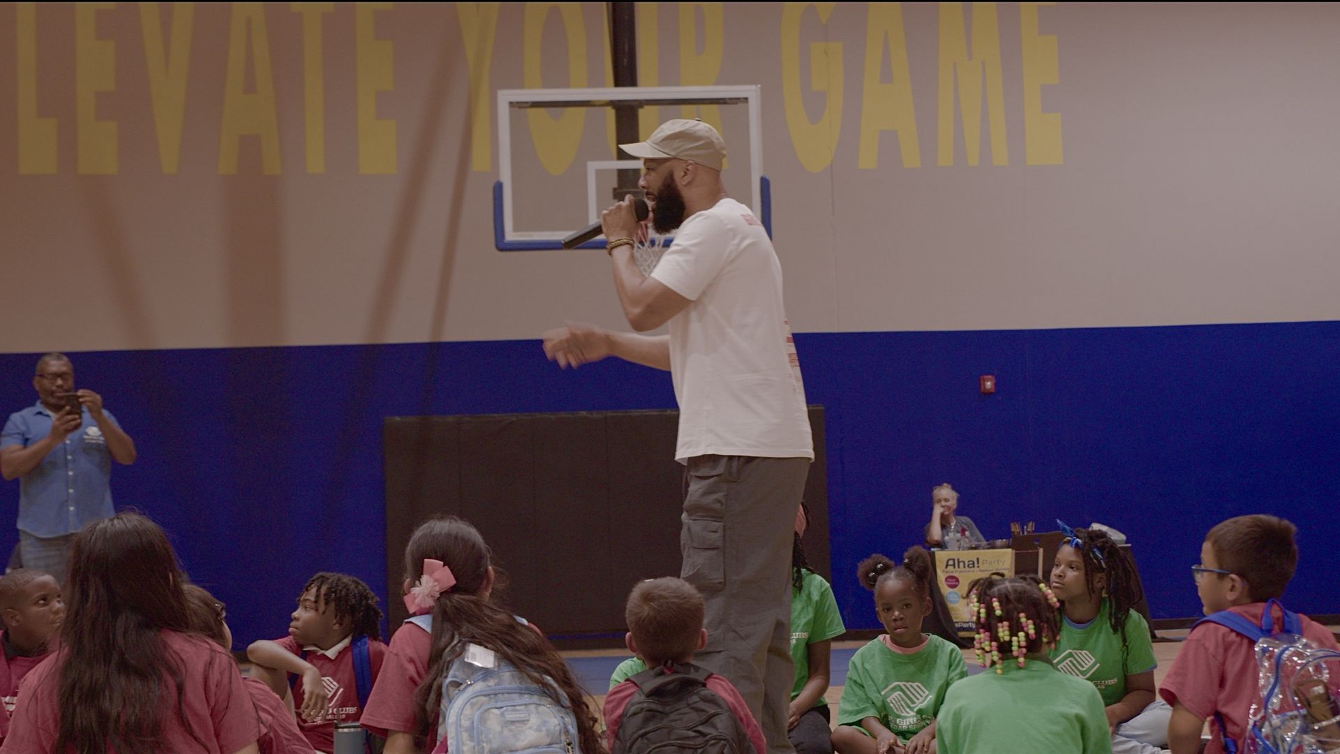 A rapper holds a microphone with children sitting around him, listening