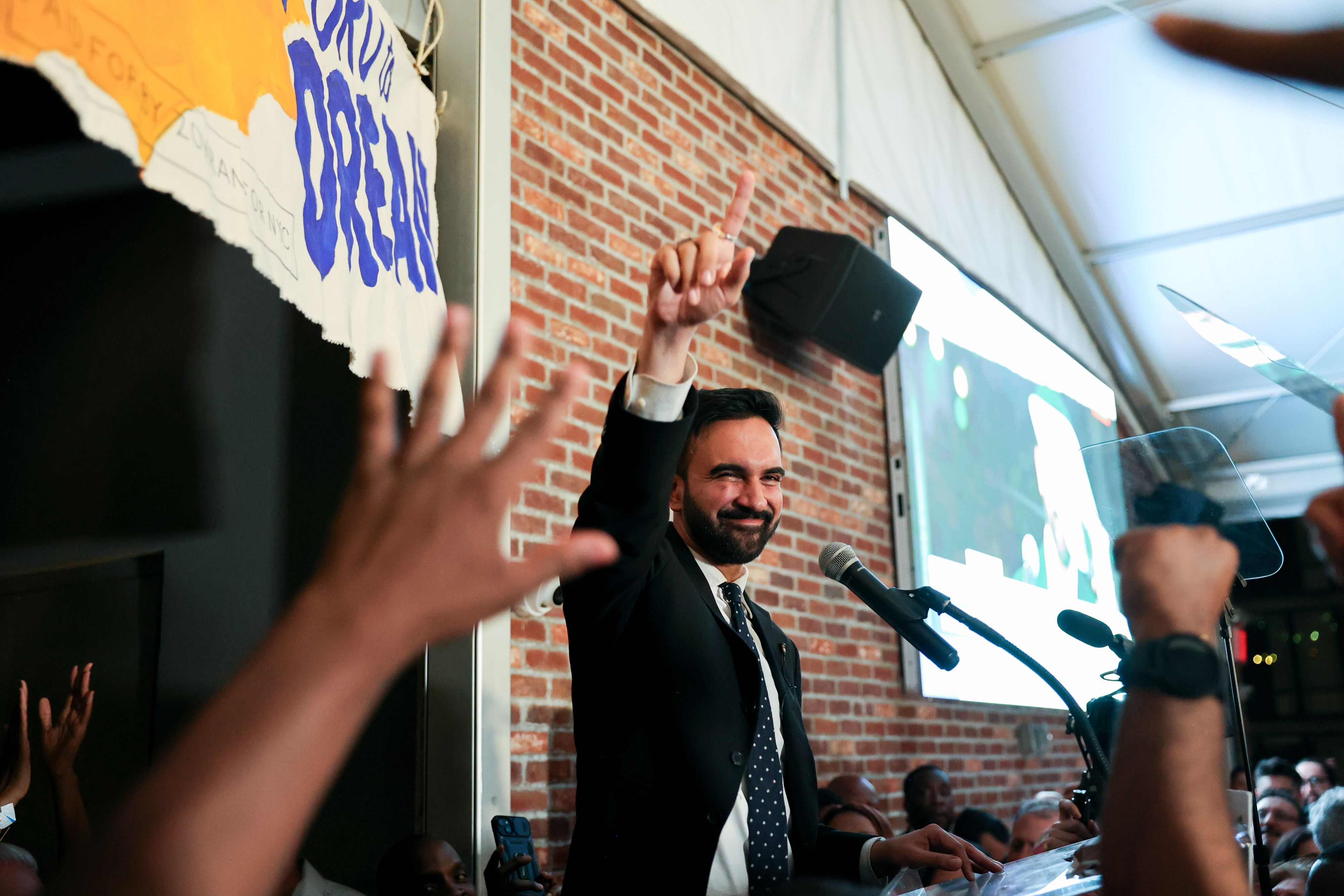A man in a suit, standing at a podium with microphones, is smiling and raising his index finger. He appears to be addressing a cheering crowd in a brick-walled venue. Hands are visible in the foreground, gesturing enthusiastically. A partially visible sign in the background reads "DREAM."