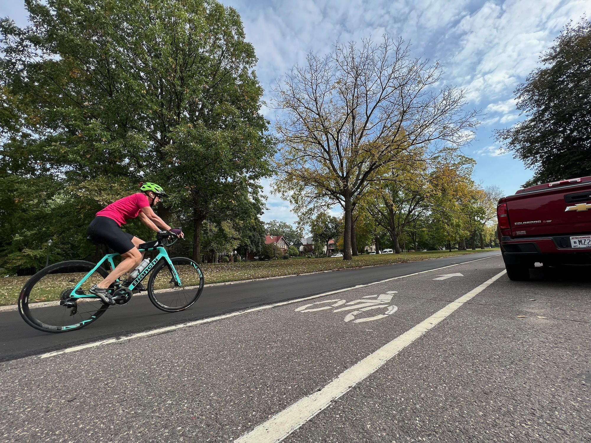 A cyclist in a pink jersey and lime helmet rides a teal road bike in a marked bike lane; a red pickup is parked on the right, with green trees and a blue, cloudy sky overhead.