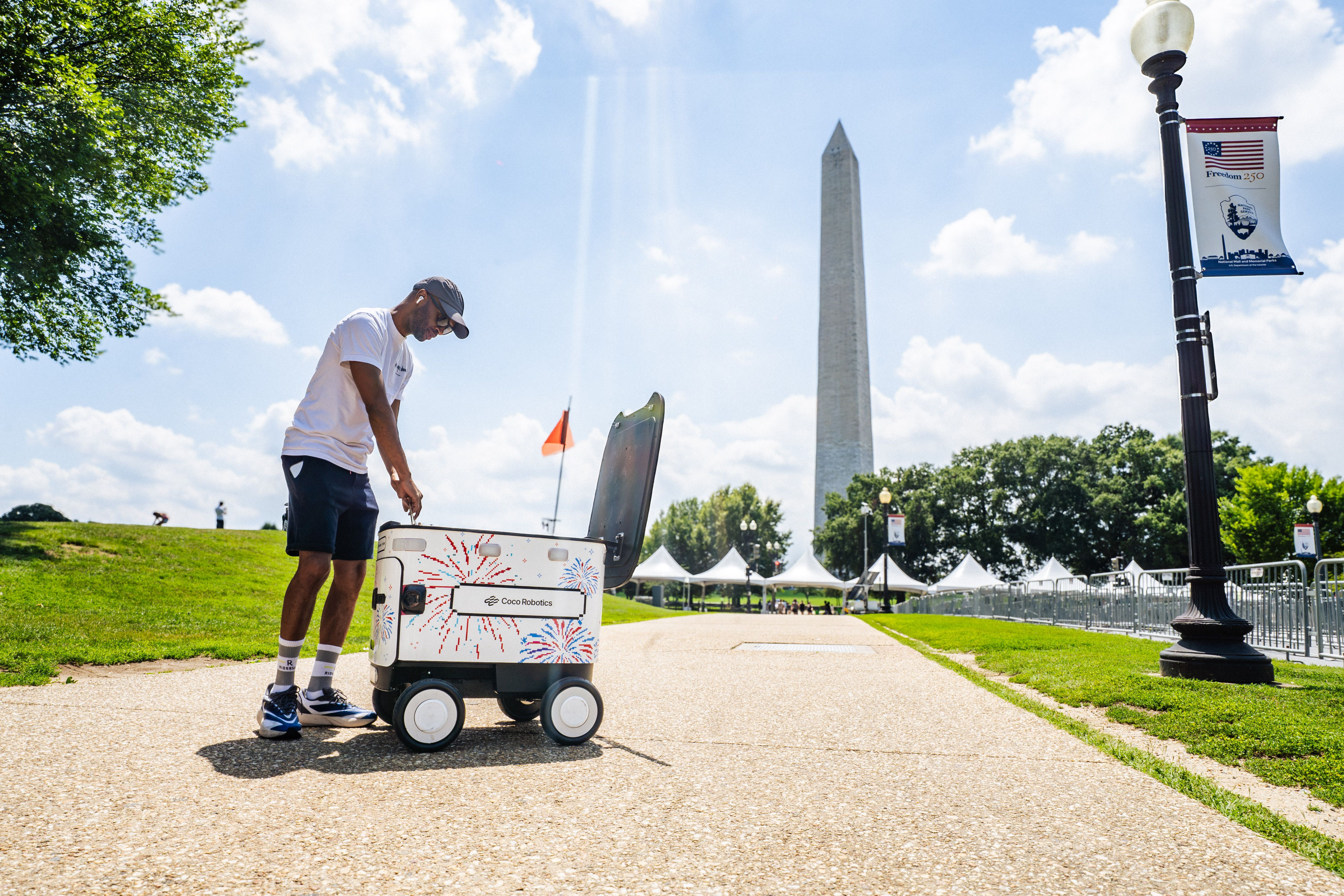 Man in a white T-shirt and cap pushes a white wheeled cooler labeled Coco Robotics, decorated with fireworks, in a sunny park with the Washington Monument in the background and tents nearby.