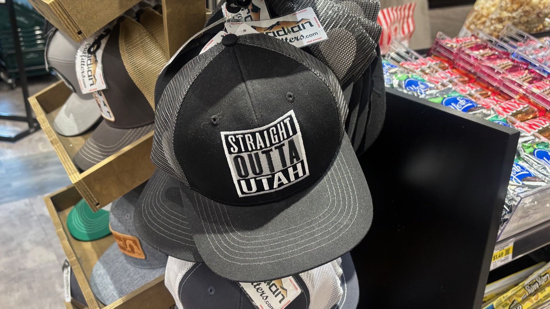 Display of various hats including a black and gray one with "STRAIGHT OUTTA UTAH" text in a store near candy shelves.