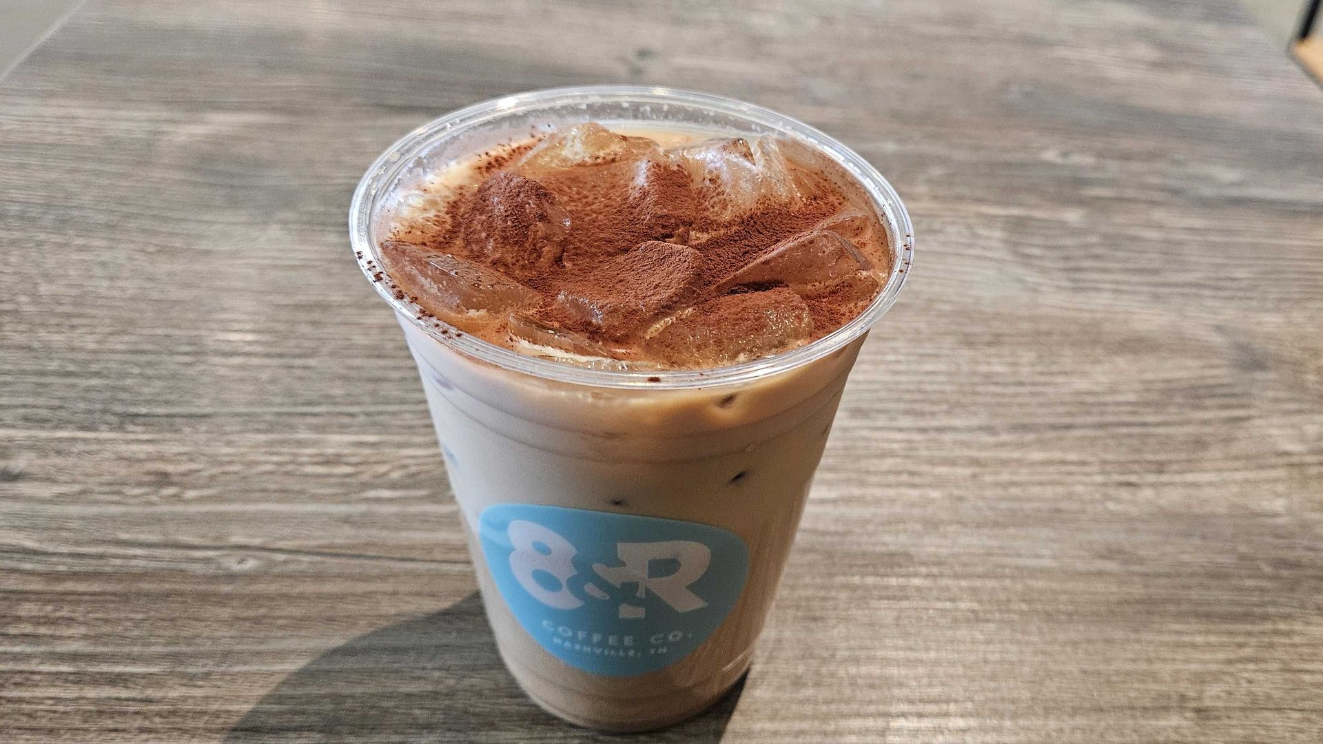 Iced coffee in a clear plastic cup with ice and cocoa powder on top, placed on a wooden table. The cup has a blue and white 8&R Coffee Co. logo.