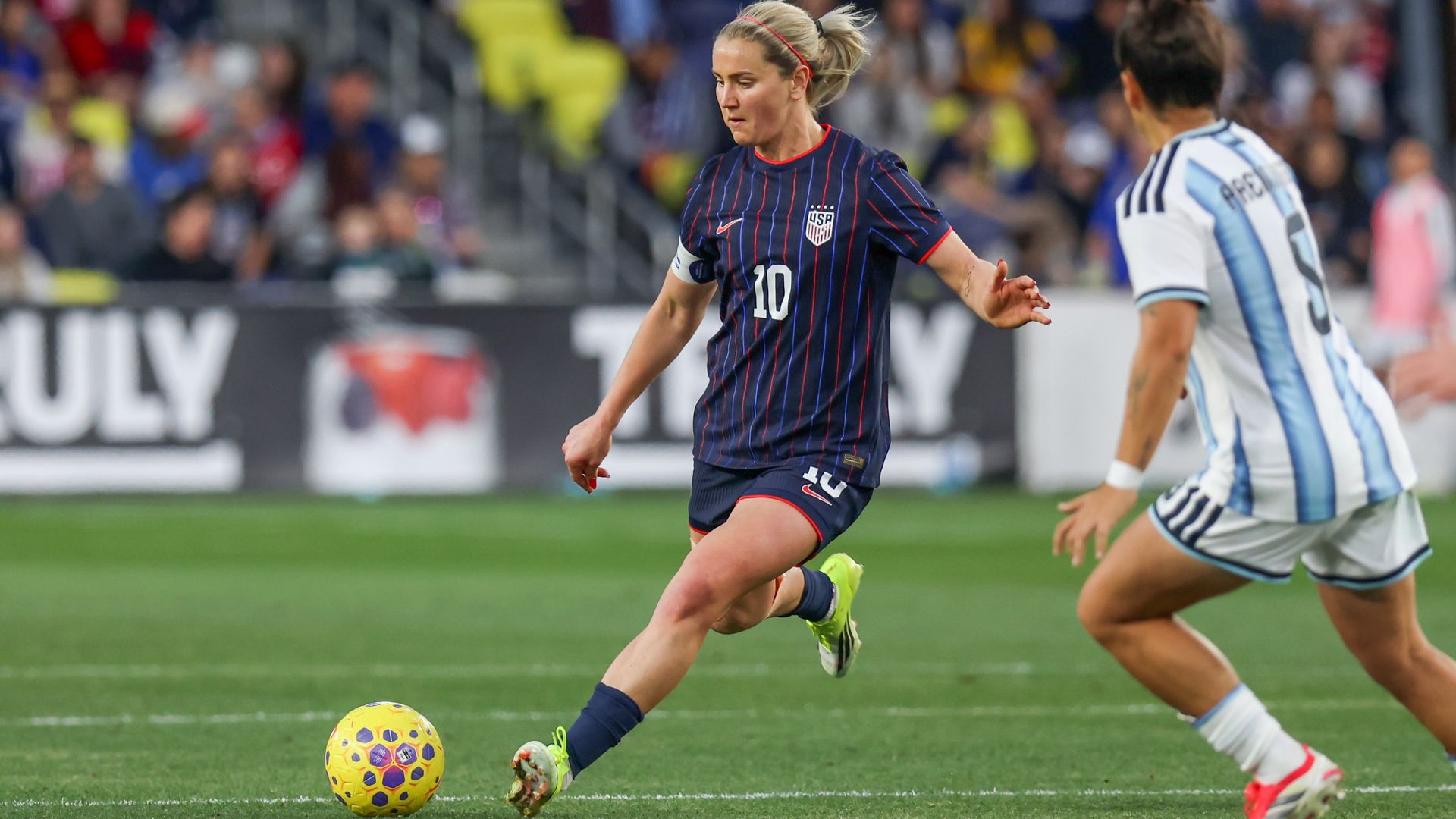 Female soccer player in a dark blue USA jersey with number 10 and red trim, blonde hair in a headband, dribbles a yellow ball on a green field while an opponent in light blue-and-white stripes closes in.