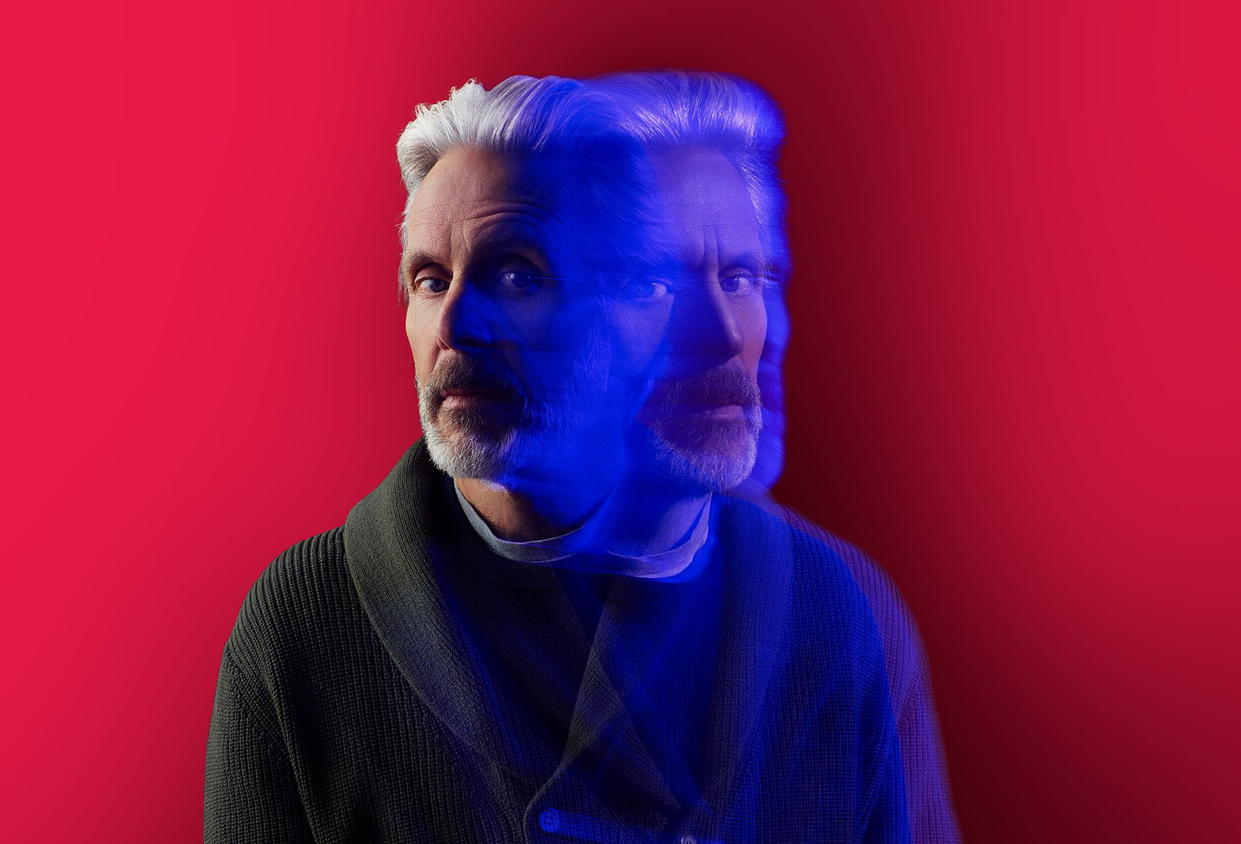 Portrait of a bearded man with gray hair in a dark textured jacket against a bright red background. A blue, semi-transparent double exposure overlays his face, creating a ghostly effect.