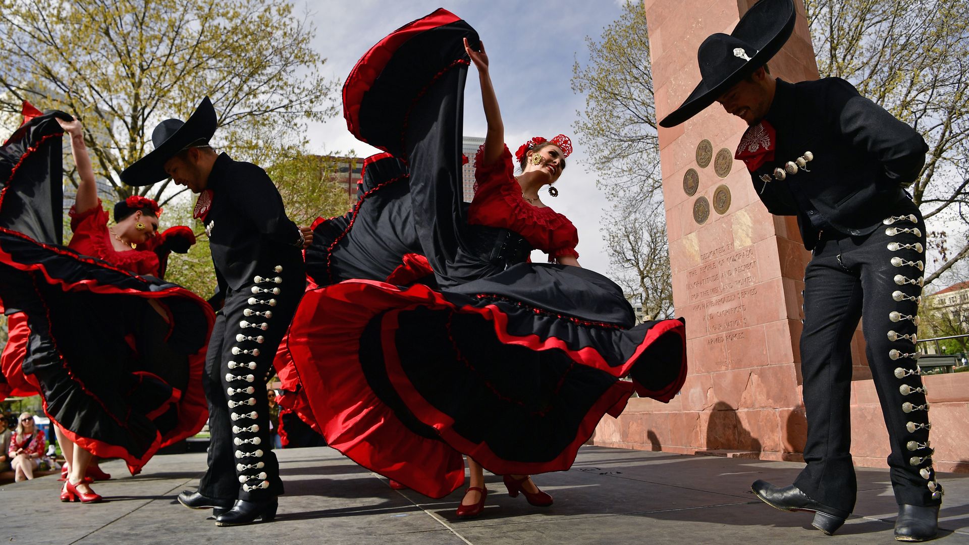 People in traditional Mexican garb dance outside in a sunny day. 
