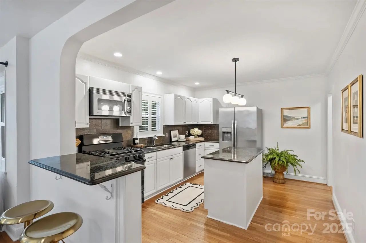 Bright kitchen with white cabinets, stainless steel appliances, granite countertops, a central island, wooden floors, and framed artwork on white walls, well-lit by ceiling lights.