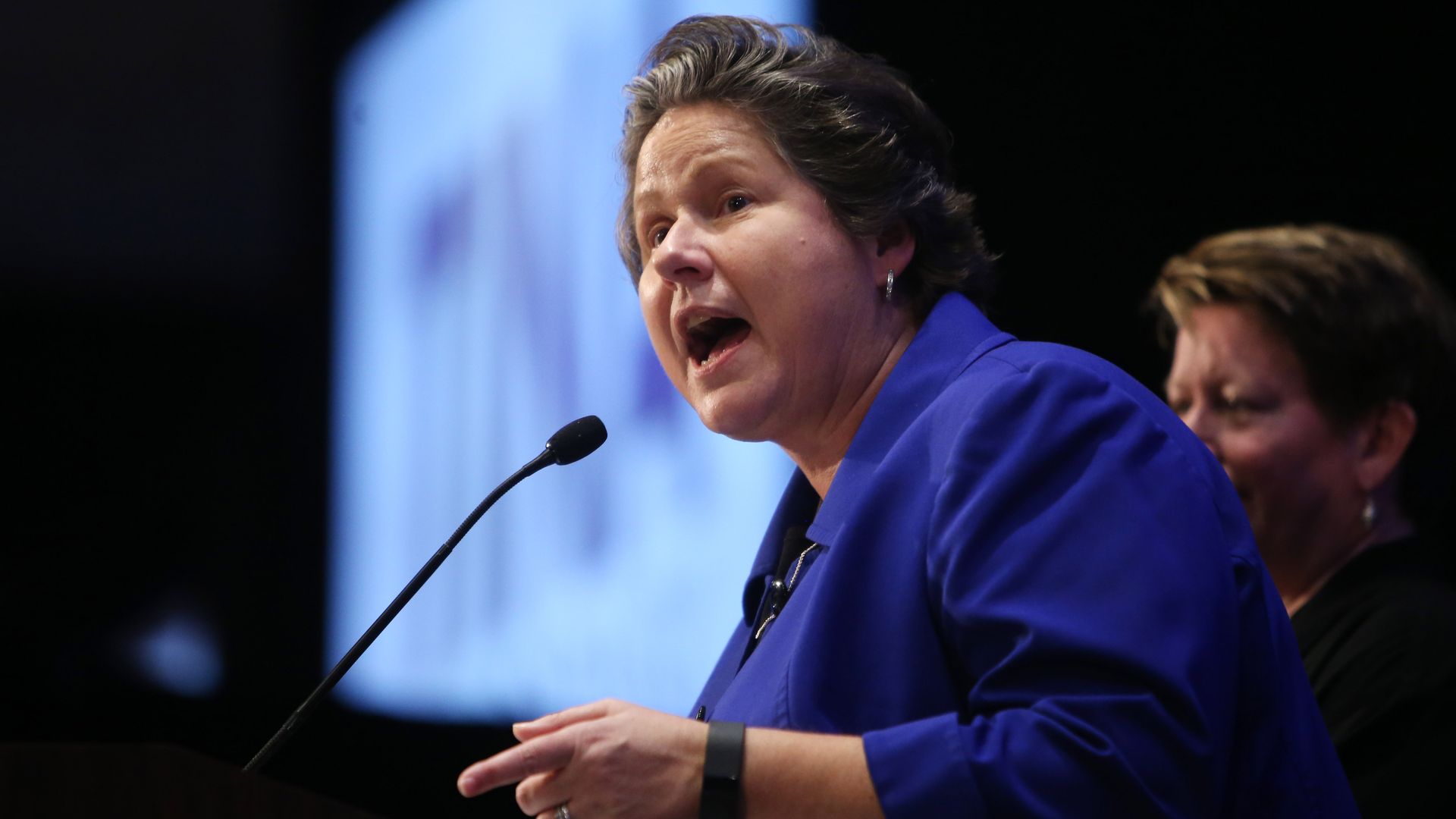 A woman in a blue suit jacket speaks at a podium into a microphone, with an indistinct image projected in the background.