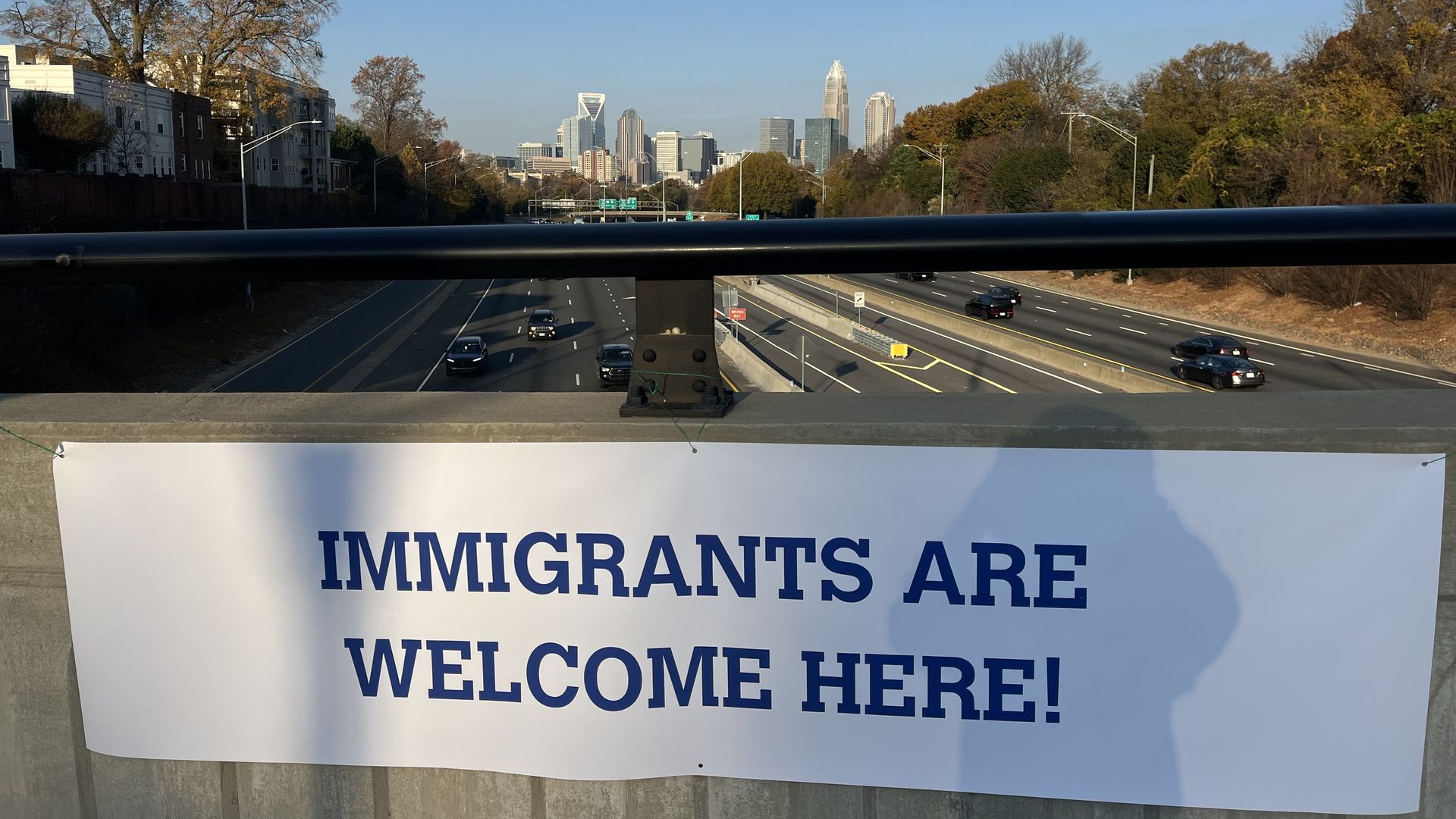 White banner on a bridge over a highway with the message "IMMIGRANTS ARE WELCOME HERE!" in blue letters, city skyline and autumn trees in the background under clear blue sky.