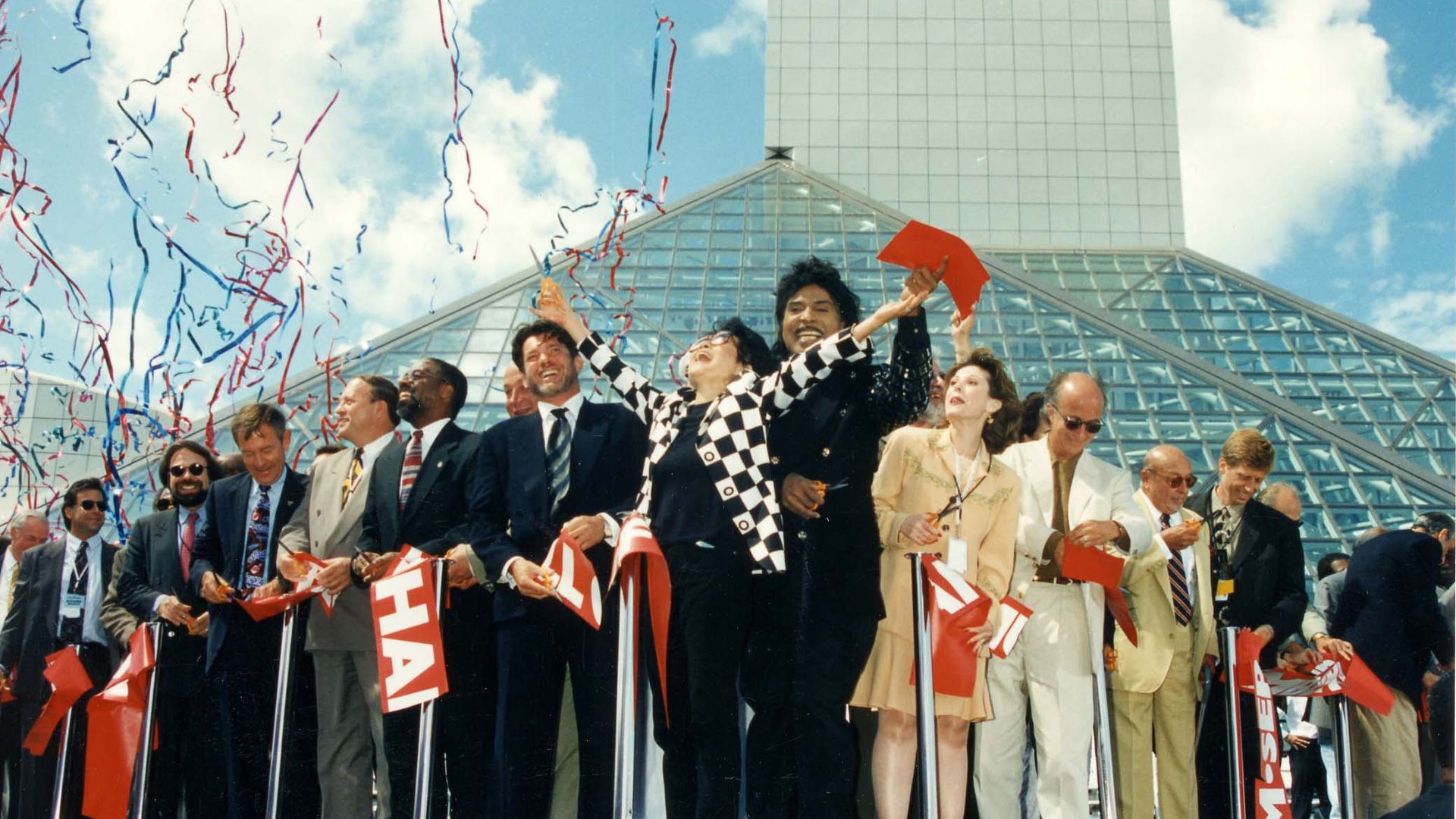 Group of diverse people in formal wear cutting a red ribbon in front of a glass pyramid building with red and blue confetti falling from the blue sky above during a celebratory event.