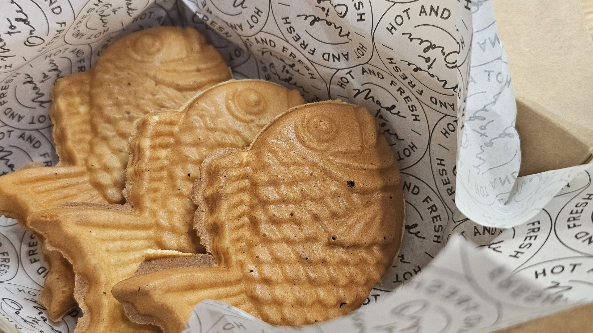Three golden-brown, fish-shaped pastries with ridged shells rest in a box lined with white paper printed with circular "HOT AND FRESH" text.