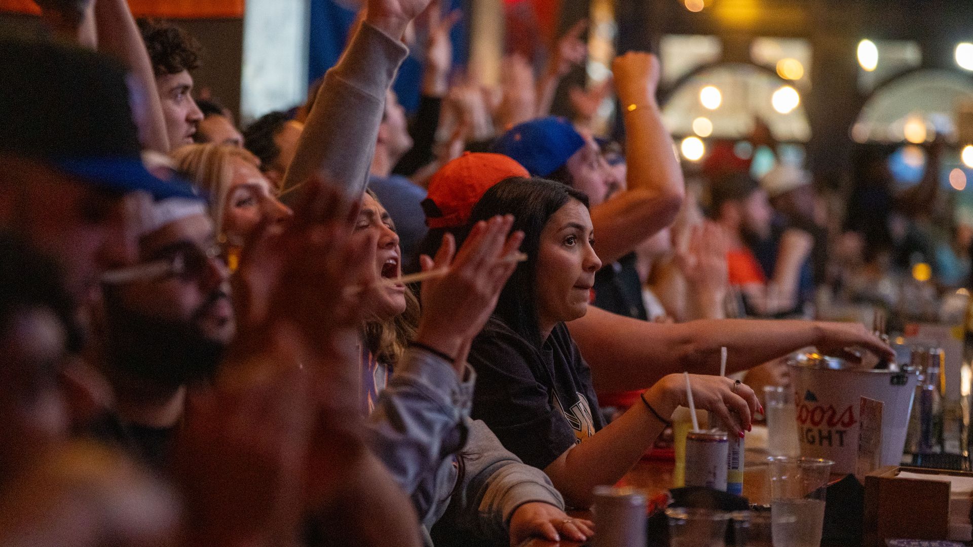 Crowded venue with fans cheering, arms raised. Foreground shows a woman with dark hair looking toward the stage; drinks and cups on the counter, warm bokeh lights in the background.