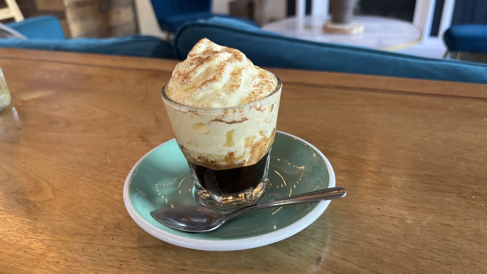 Small glass of coffee topped with egg custart cream and a sprinkle of nutmeg, placed on a teal saucer with a silver spoon on a wooden table in a cozy cafe setting.