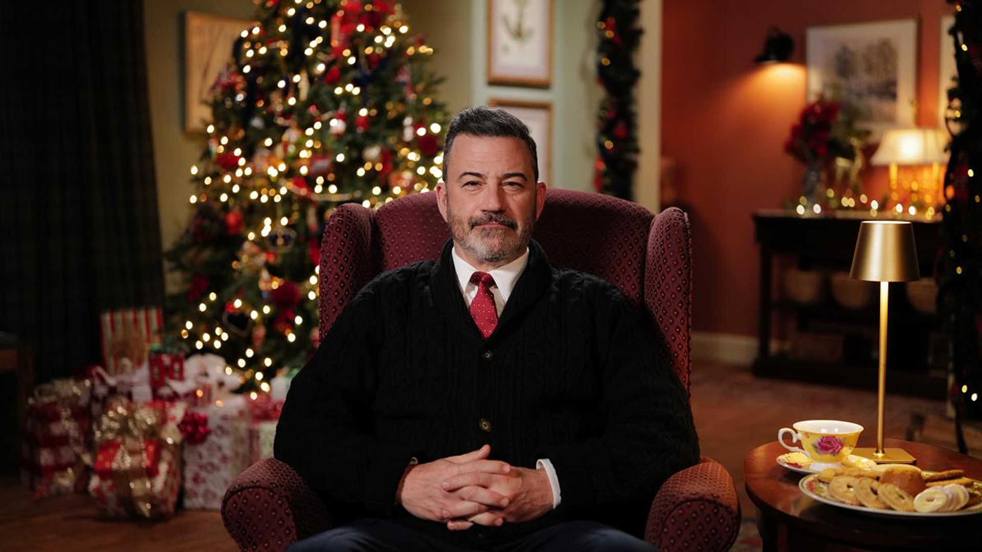 A fair-skinned, graying, dark-haired and bearded, dark brown-eyed talk show host Jimmy Kimmel looks into the camera while wearing a black buttoned-up cardigan, white shirt and red tie, hands clasped, and sitting in a red armchair beside cookies, a tea cup in front of a Christmas tree with lights.