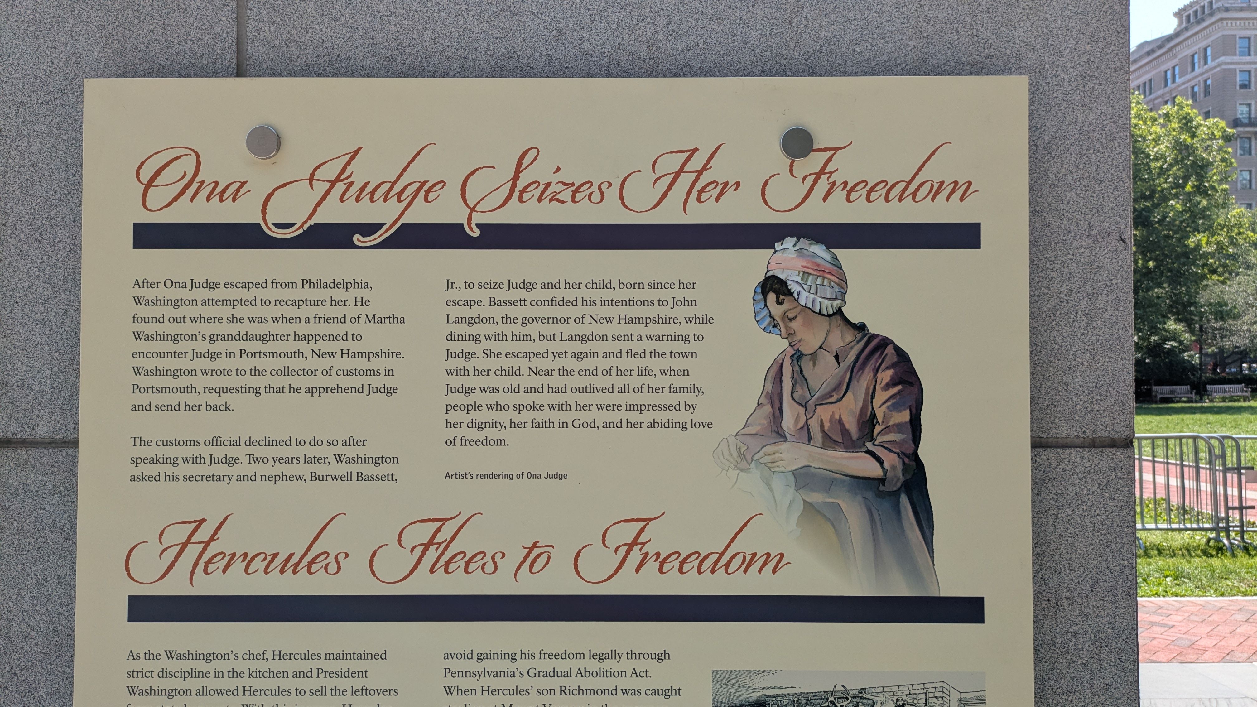 Large beige outdoor exhibit board reading "Ona Judge Seizes Her Freedom" in red script, with a painting of a woman in a bonnet on the right, and a dark blue underline, set beside a granite pillar.
