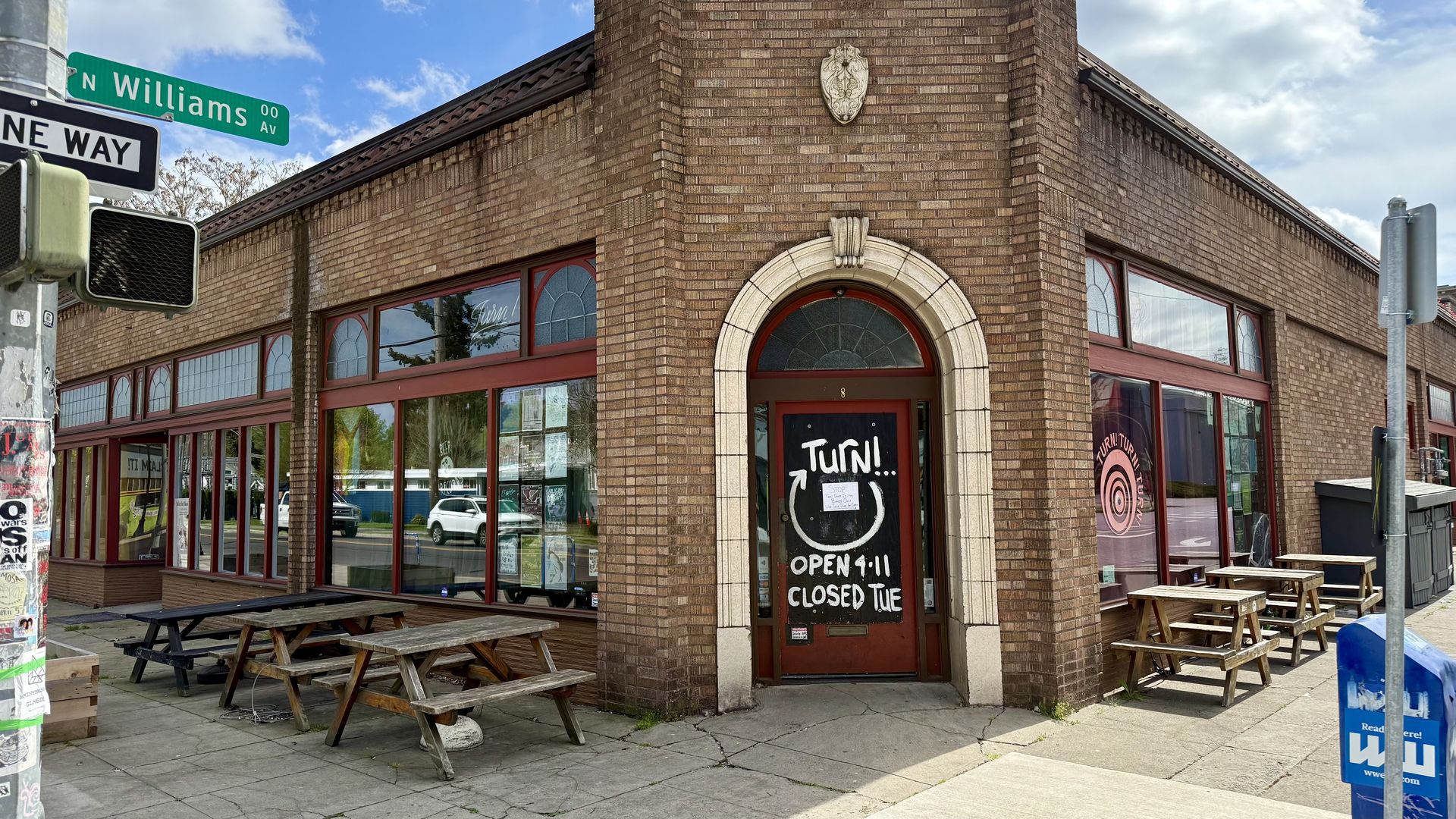 Brick corner building with red-framed windows and a large light-stone arched doorway; crest above; outdoor wooden picnic tables; street signs reading "N Williams Ave" and "One Way" under a blue sky.