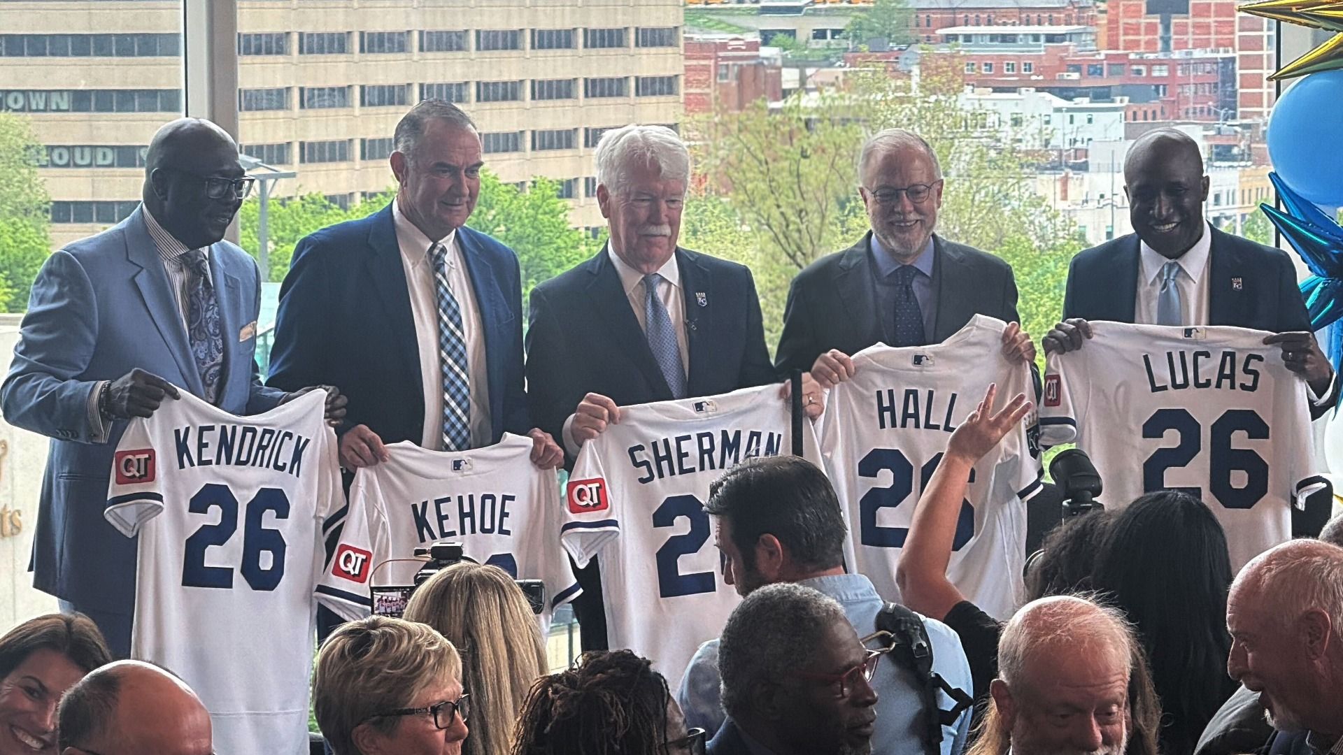 Five men in suits hold up white baseball jerseys with names Kendrick, Kehoe, Sherman, Hall, Lucas; blue balloons decorate the right, city skyline in the background, crowd watching.