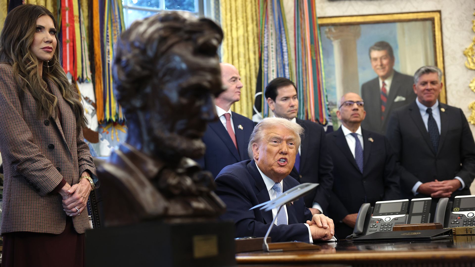 Donald Trump sitting at a desk flanked by Kristi Noem, Marco Rubio and others. A statue of Abraham Lincoln is in the foreground 