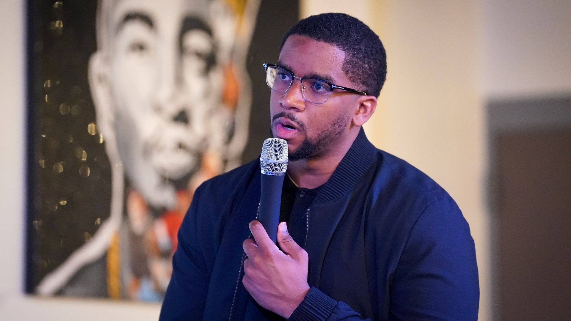 A man with a beard, wearing a dark jacket, speaking into a microphone indoors with a colorful, blurred portrait painting in the background.