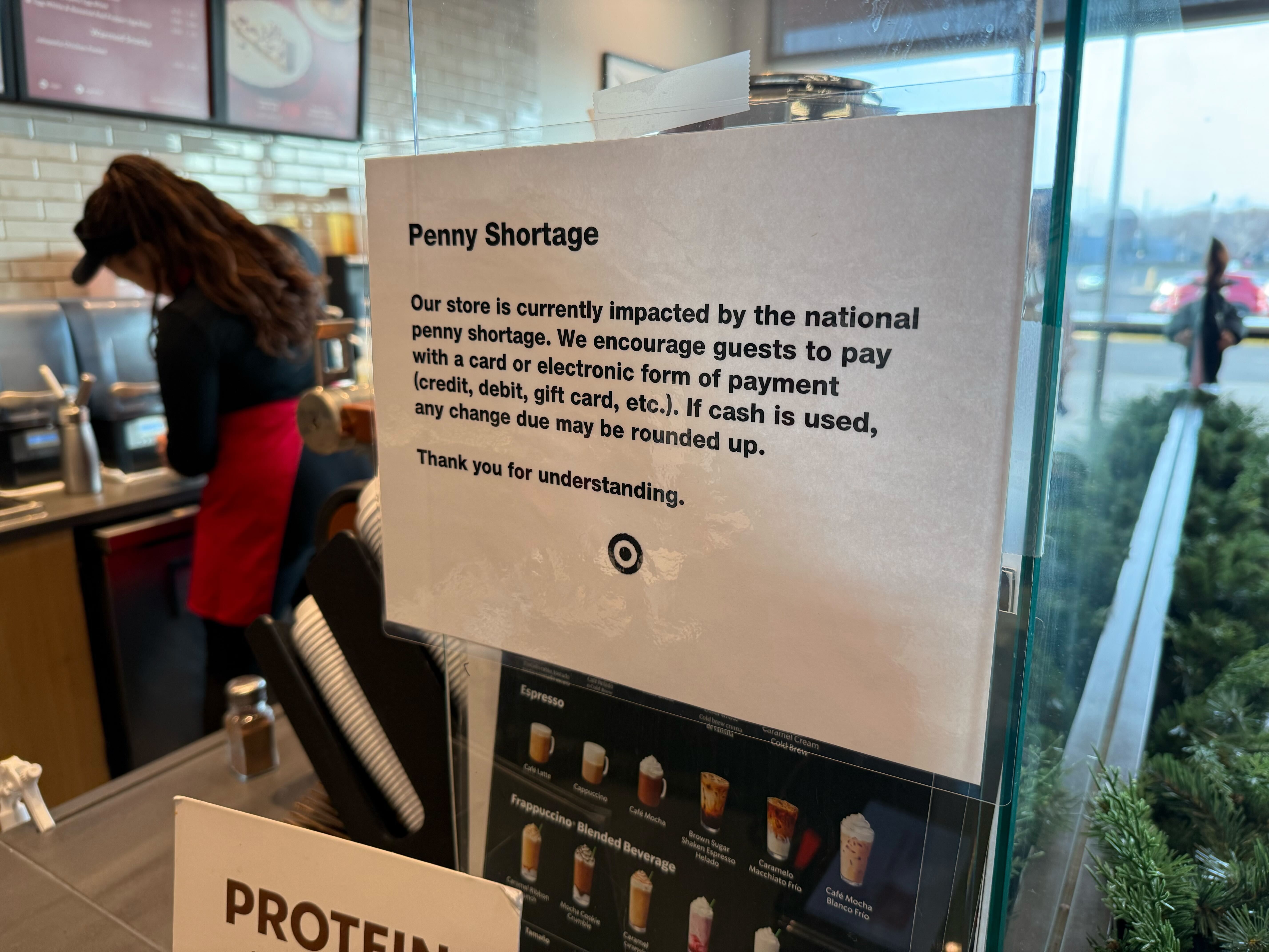 Sign at Starbucks in a Target explains the penny shortage, encouraging electronic or card payments, with cash change possibly rounded up.