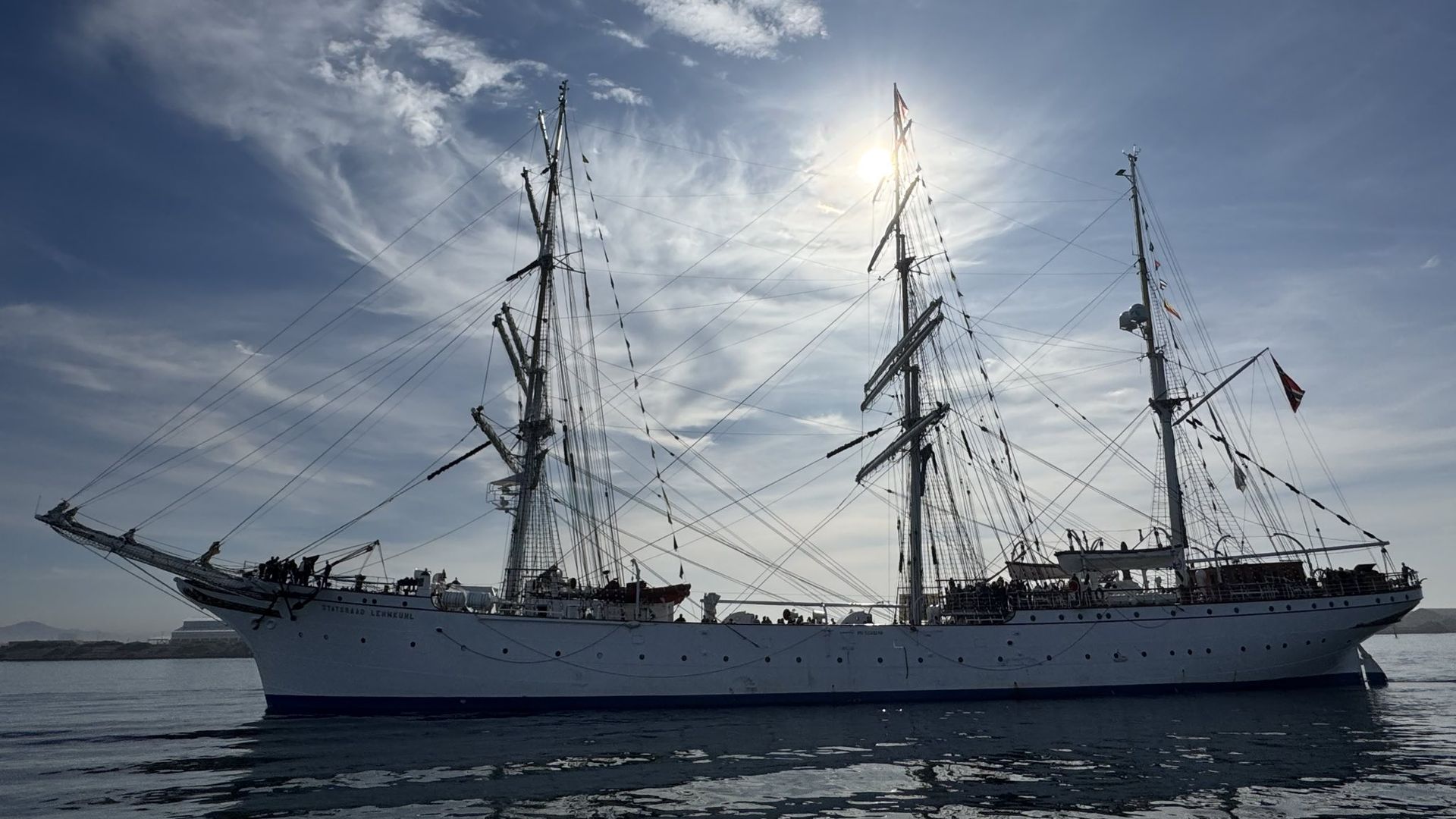 Large white sailing ship with three masts and rigging, floating on calm water under a partly cloudy blue sky with the sun shining behind the central mast.
