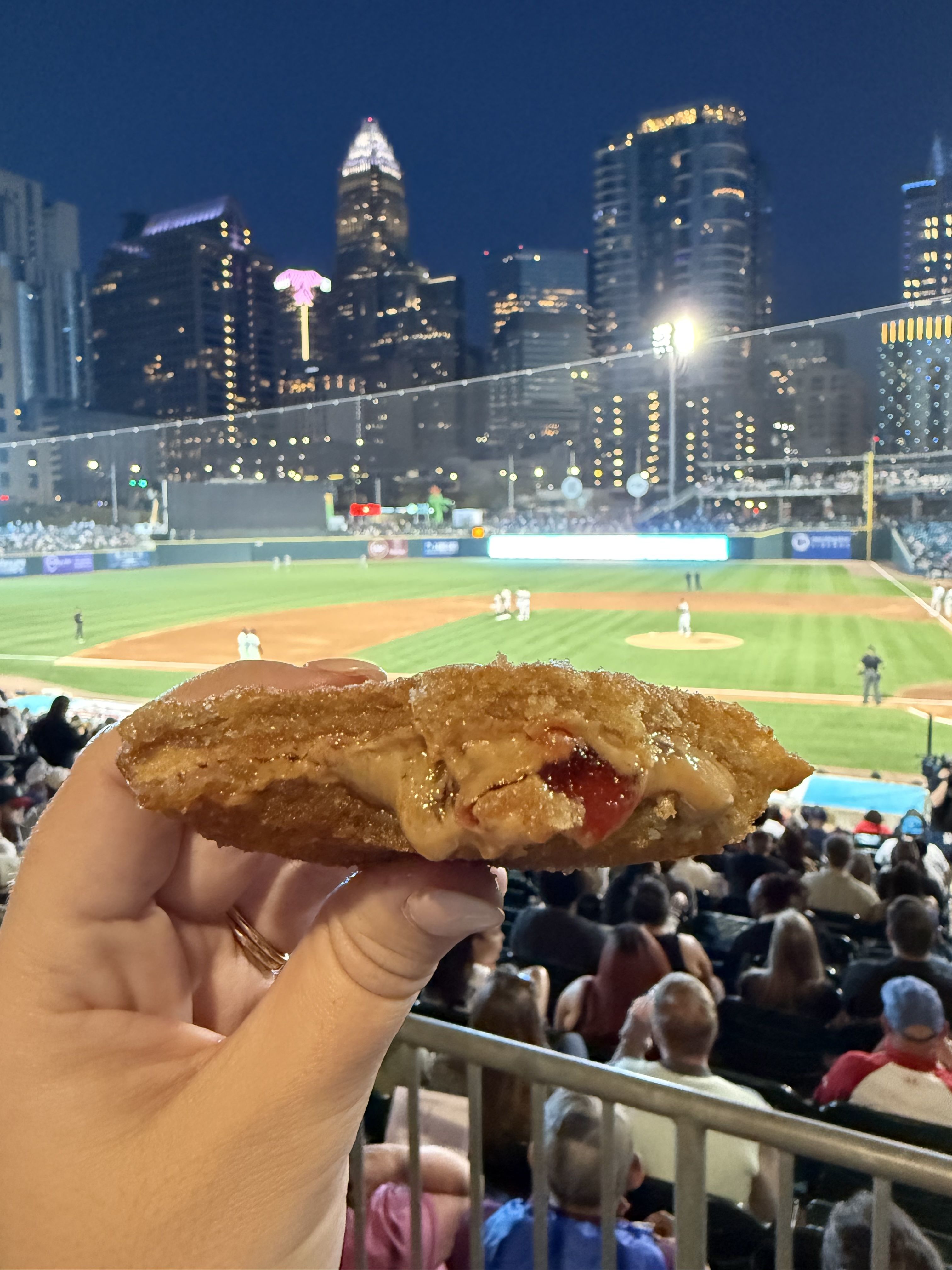 A hand holds a gooey, deep-fried peanut butter and jelly sandwich with the stadium crowd and Charlotte skyline glowing in the background.