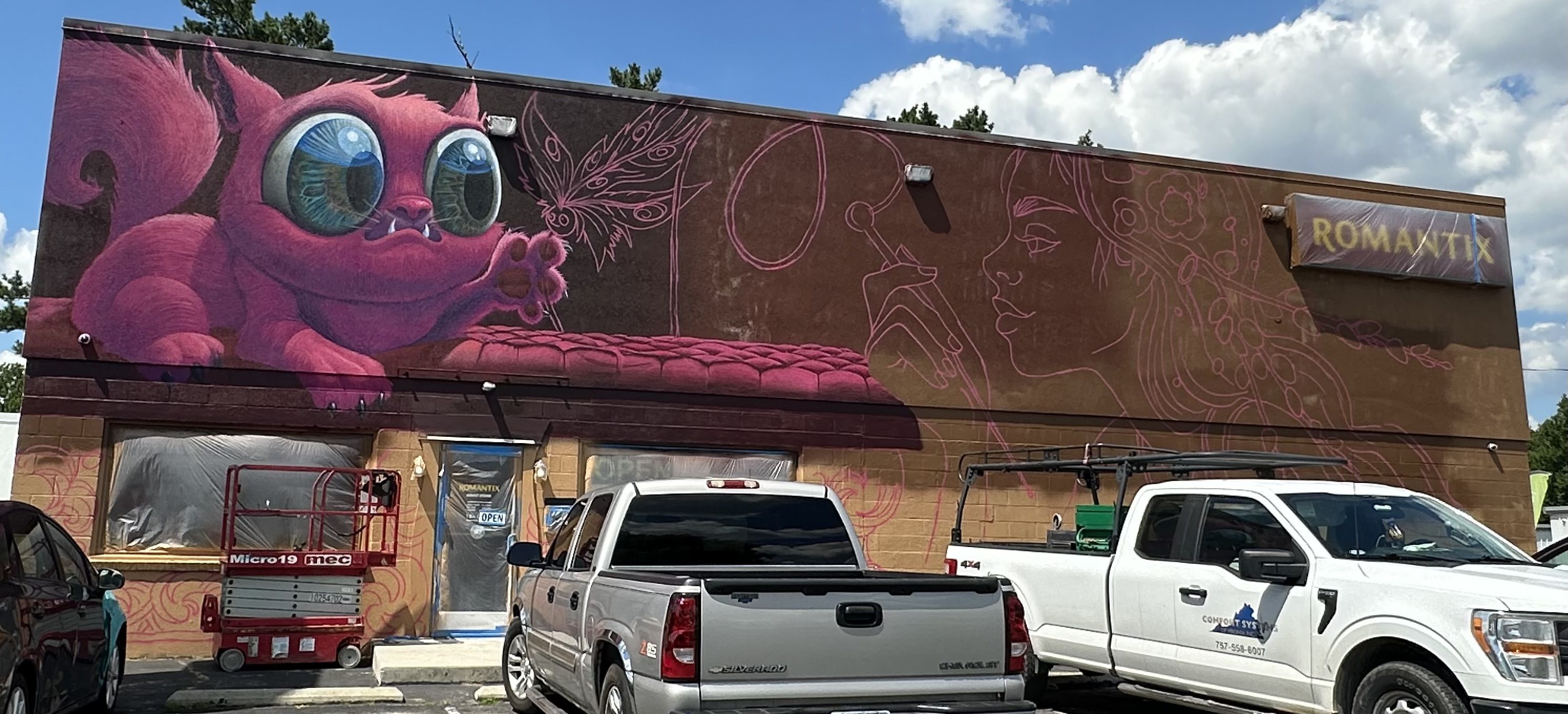 Colorful mural on a building shows a large pink creature with blue eyes, pink line-art figures across the wall, a ROMANTIX sign on the right, and white pickup trucks in front.