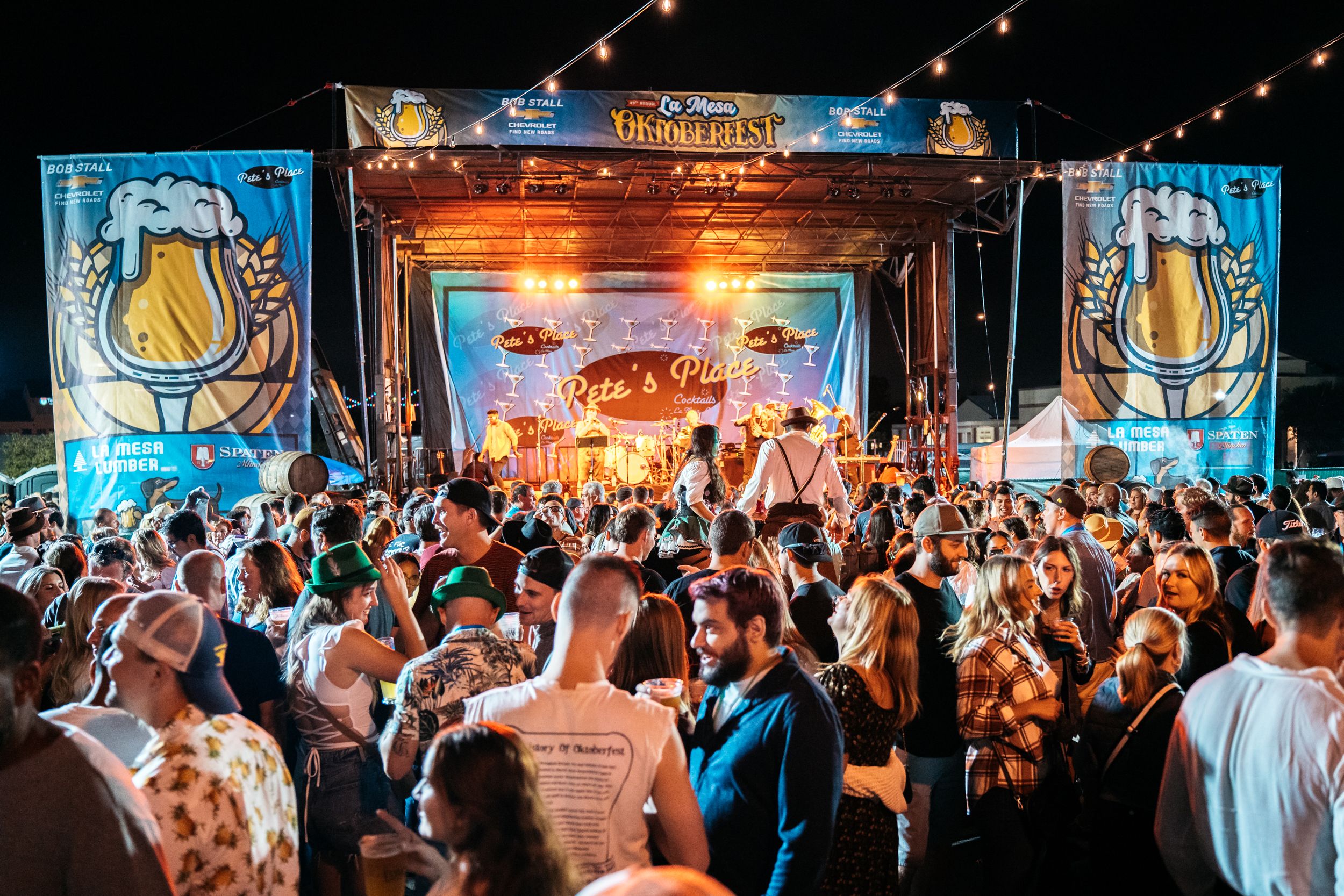 Night festival scene with a crowded beer-stage, banners reading "La Mesa Oktoberfest" and beer-mug graphics. String lights glow above as a band performs and the crowd cheers.