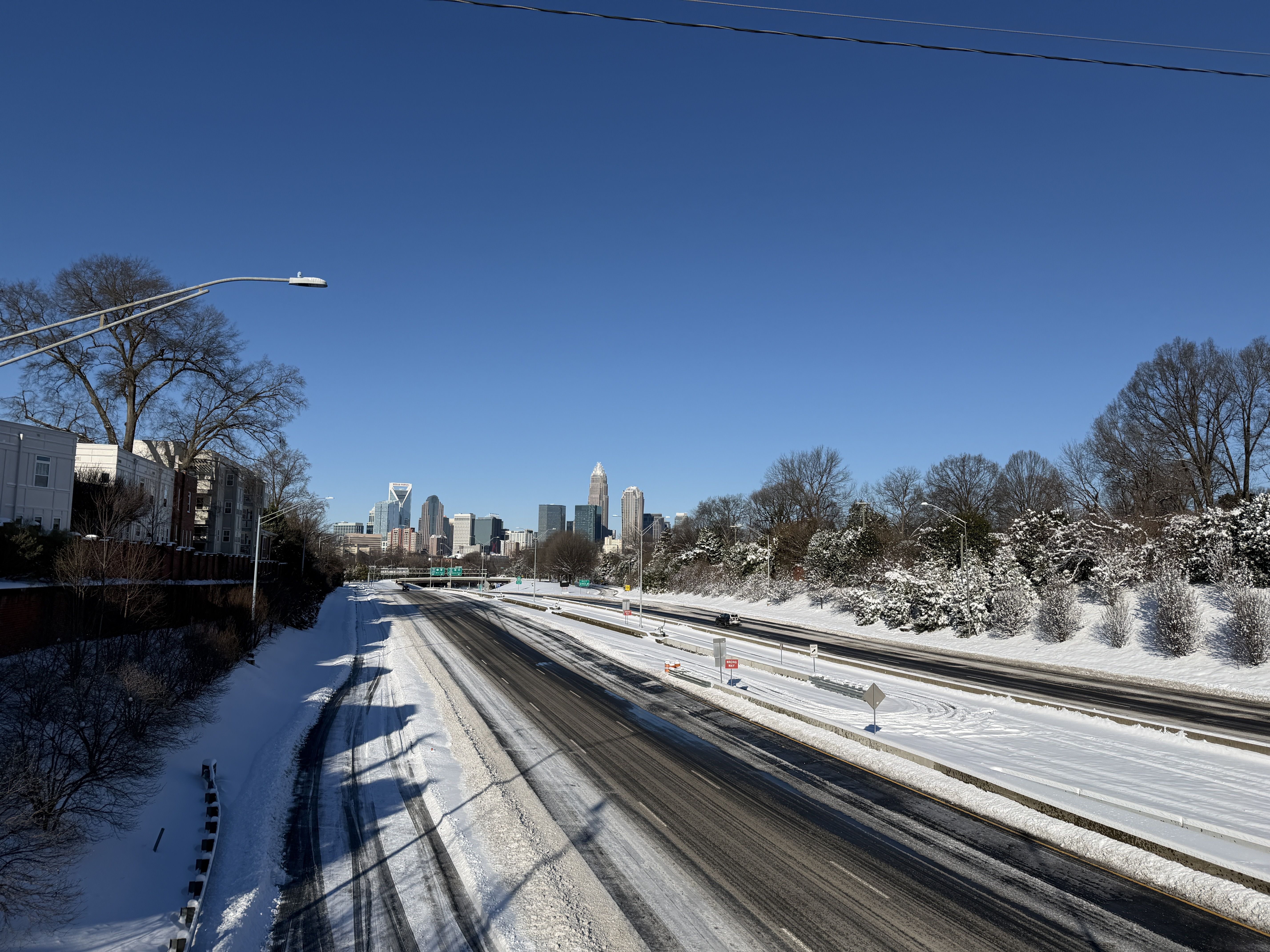 Snow-covered highway with light traffic, bare trees, and clear blue sky, with a city skyline in the distance under bright daylight.