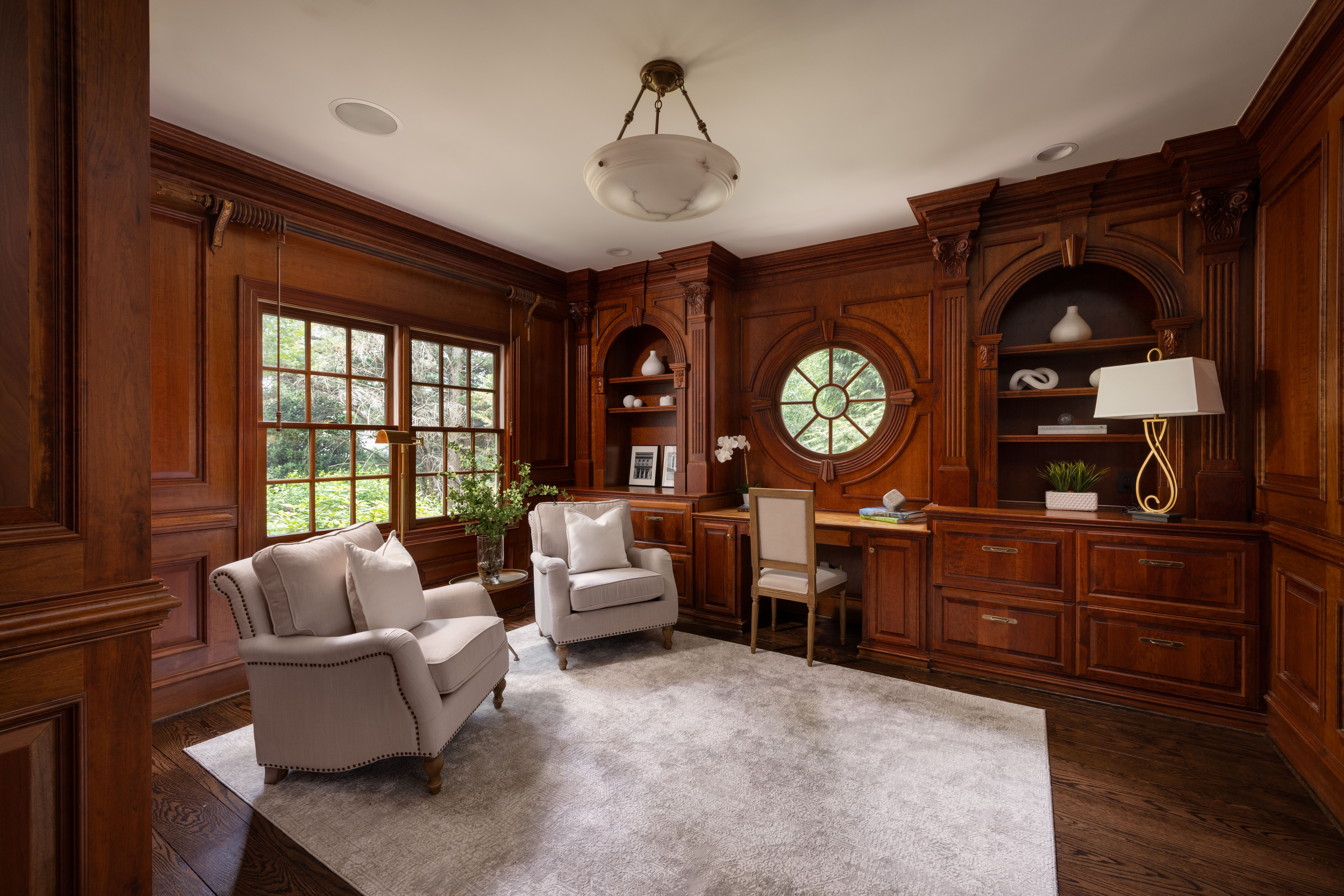 Luxurious wood-paneled study with ornate built-in shelves, arched and circular windows, and a central brass ceiling light. Cream armchairs, a desk, and greenery outside.