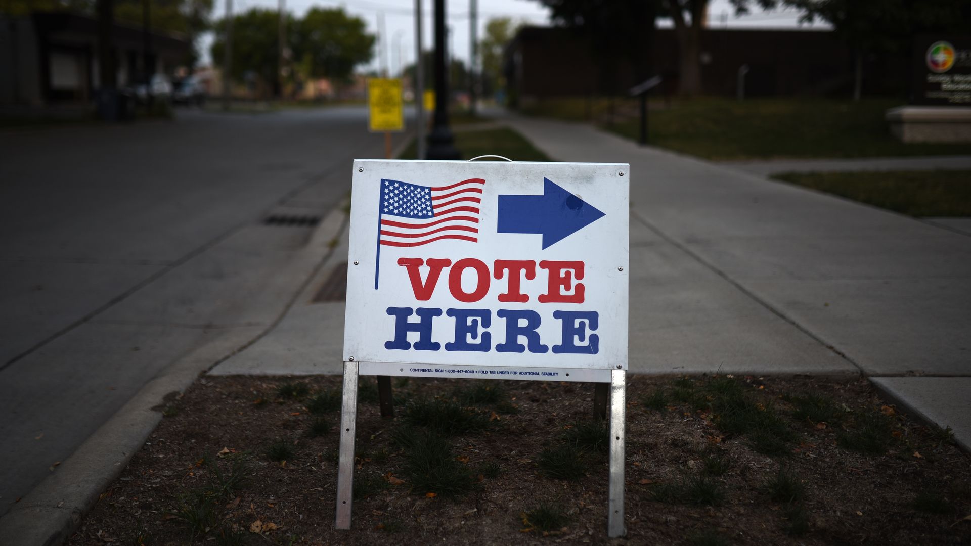 "Vote Here" sign with American flag on a sidewalk near a polling station