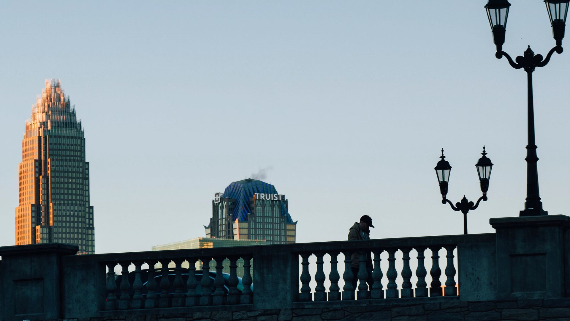Silhouette of a person walking on a stone bridge with ornate lampposts, against a clear sky and two tall city buildings, one labeled "Truist," during sunset.
