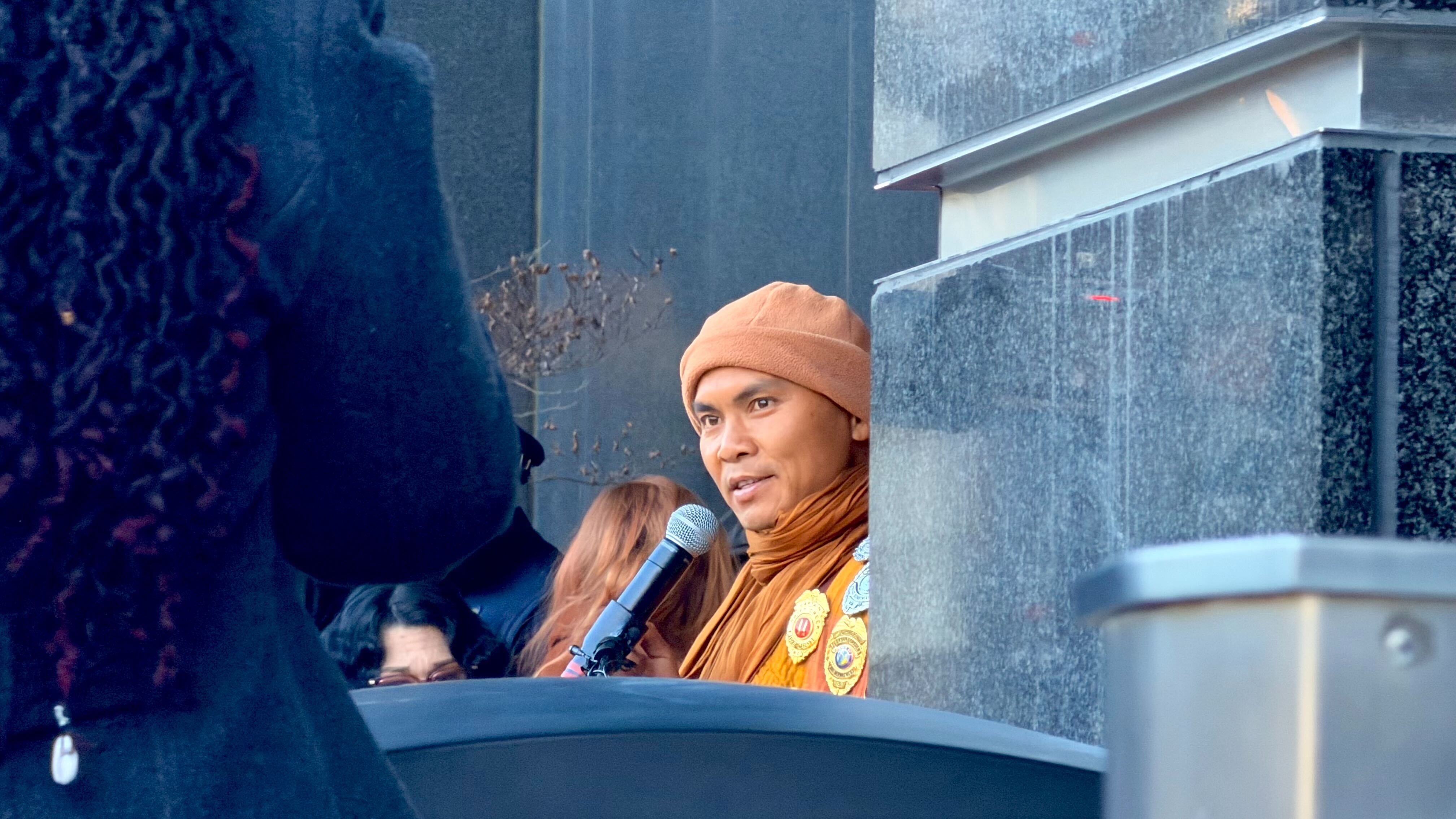 A person wearing an orange hat and matching scarf speaks into a microphone near a gray stone structure with a flame visible at the top right.