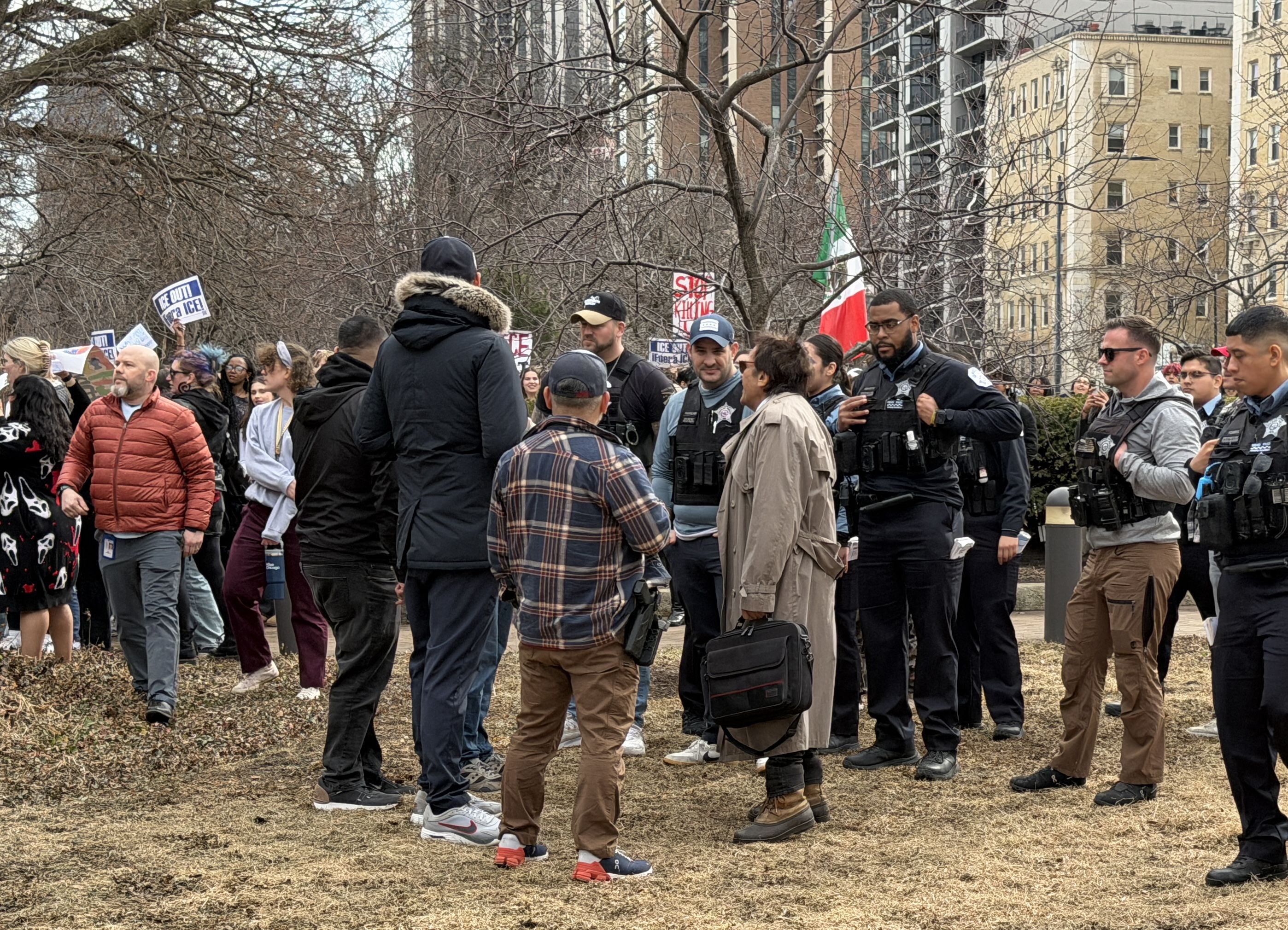 Group of protesters holding signs like "ICE OUT! Free Our ICE" and police officers in black uniforms with gear, standing on dry grass with city buildings and leafless trees in background.