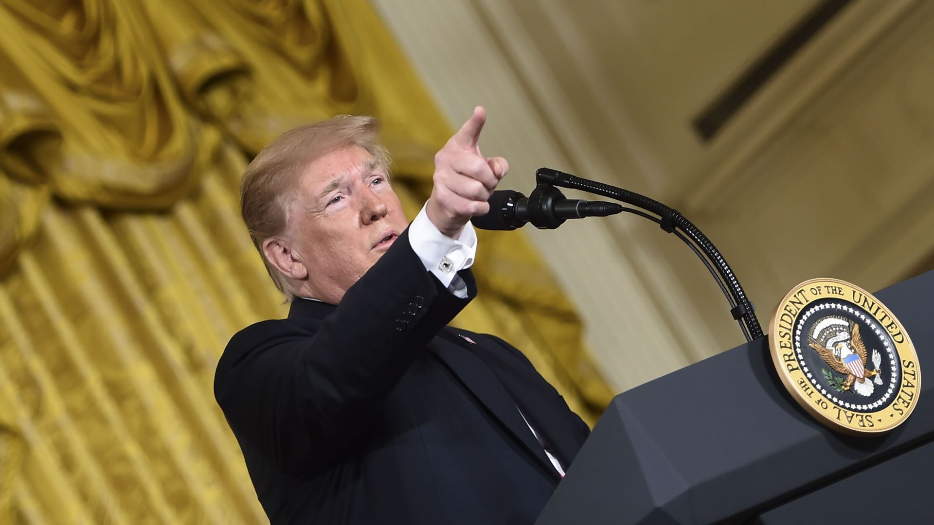 Donald Trump points from a podium before a gold drape.