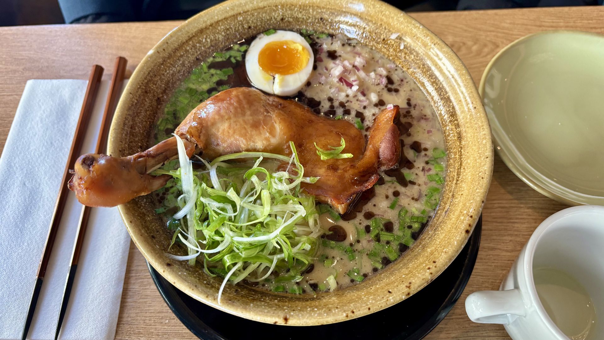 Top-down view of a ramen bowl with a roasted chicken leg, a halved soft-boiled egg, chopped green onions, and sesame seeds in a creamy broth; chopsticks and napkin to the left.