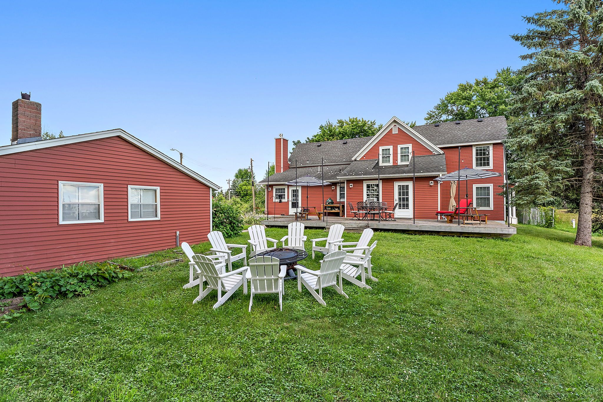 Red two-story house and detached garage with white-trimmed windows, surrounded by green lawn. White Adirondack chairs arranged around a fire pit in the yard under a clear blue sky.