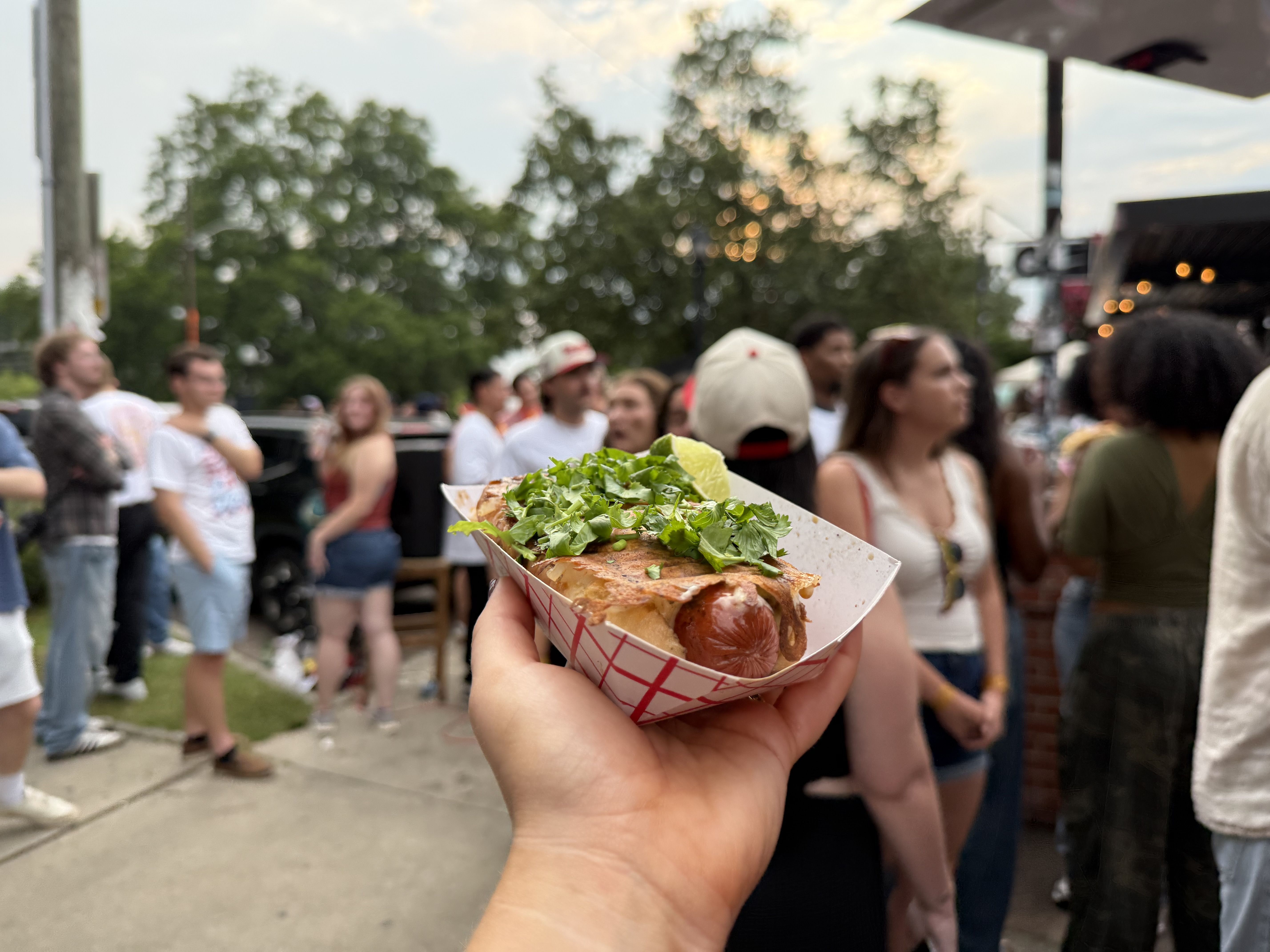 A hand holds a paper tray with a cilantro-topped hot dog and a lime wedge, foreground; a crowd and trees blur in a lively outdoor festival in the background.