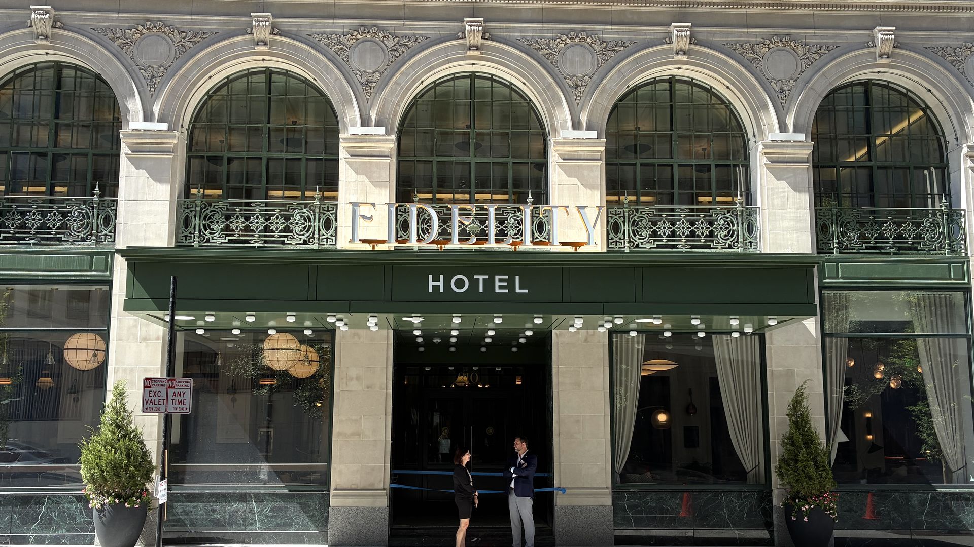 Facade of a grand hotel with five large arched windows and decorative iron balconies. A green awning with HOTEL spans the entrance; two people speak outside among potted plants.