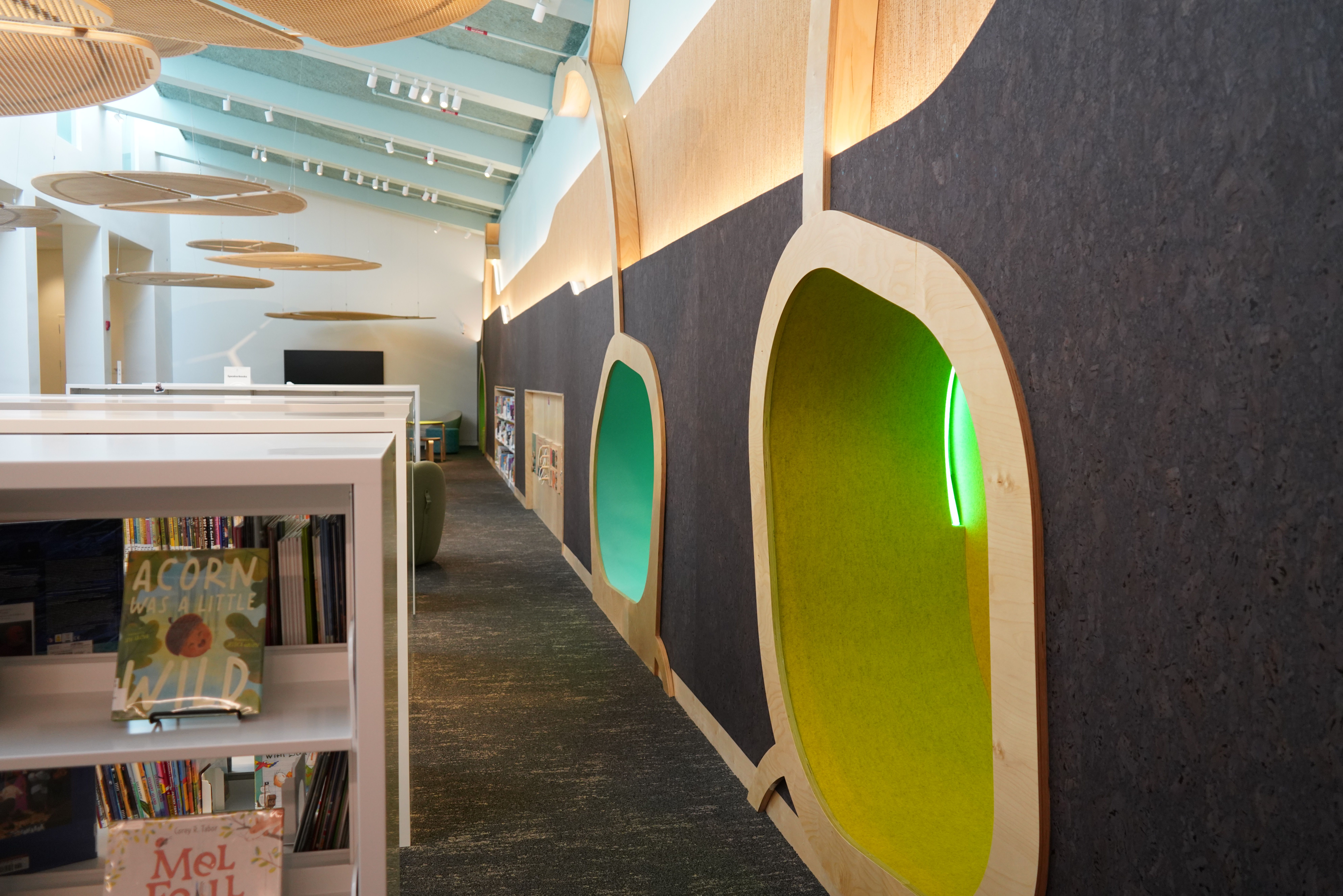 Interior library or reading space with white bookshelves in the foreground, circular wood-cut wall panels with green and yellow centers along a dark textured wall, and hanging wooden discs under a pale blue ceiling.