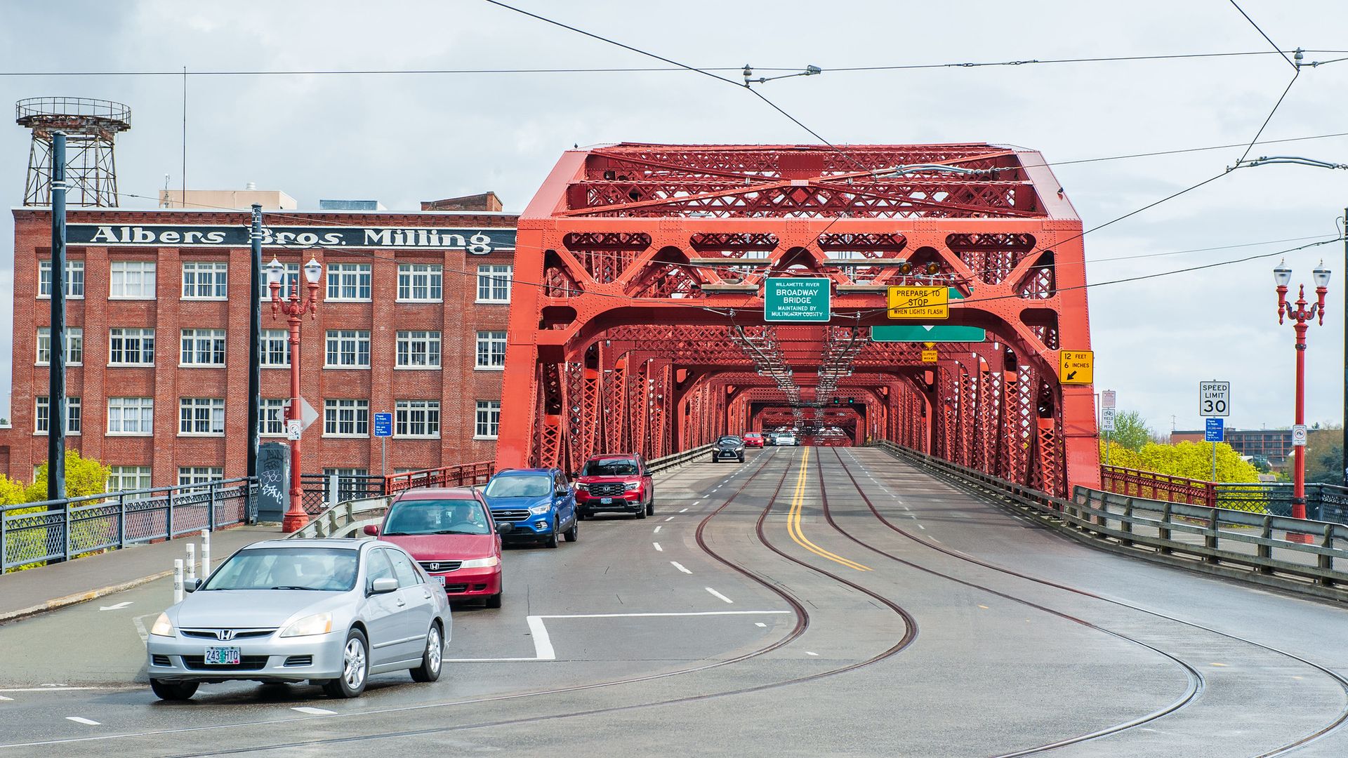 Red iron truss bridge with tram lines and overhead wires spans a street; cars drive across while a brick "Albers Bros. Milling" building sits on the left; cloudy sky overhead.