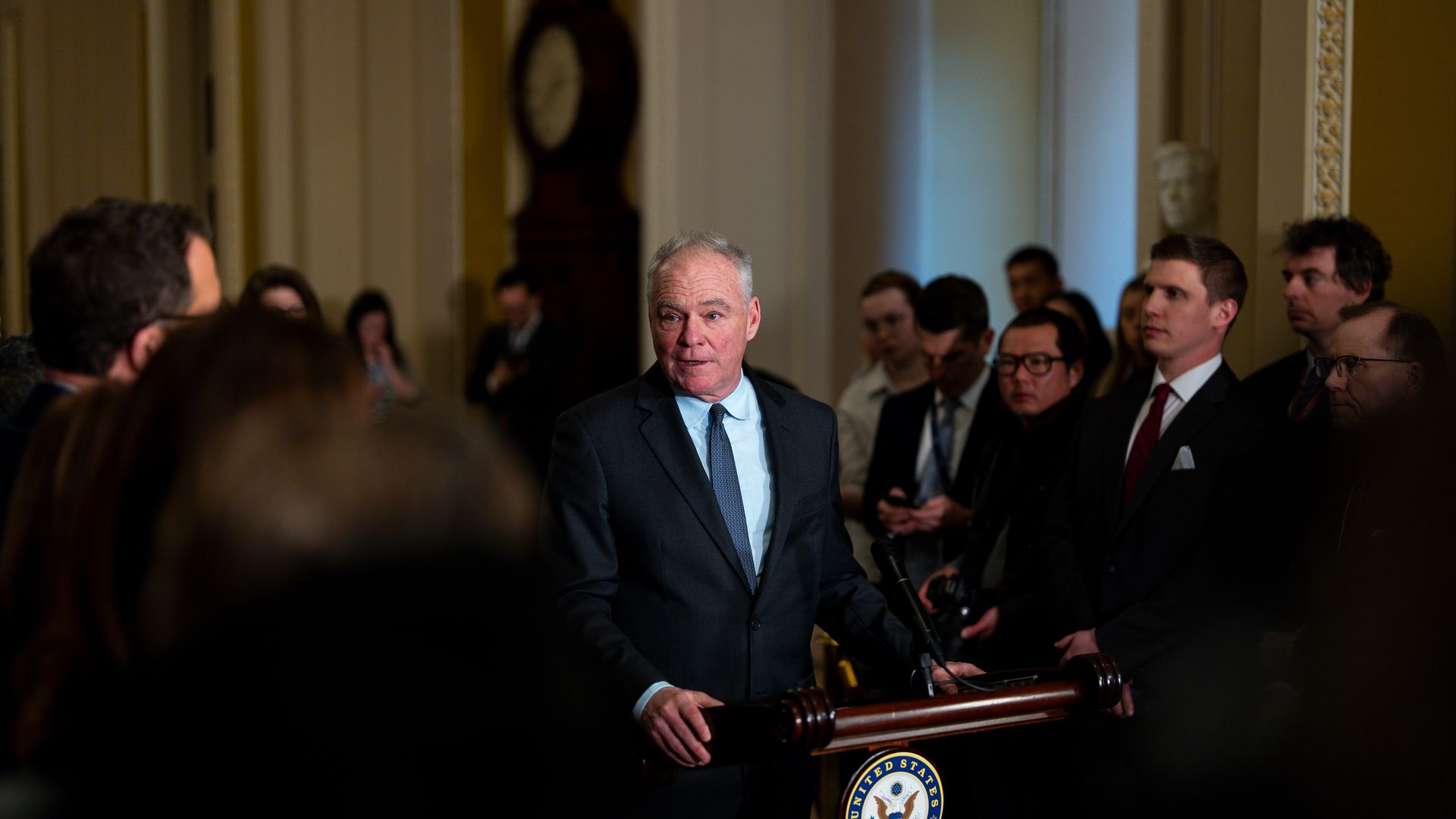 Man in a dark suit speaks at a United States Senate podium surrounded by a attentive crowd in a formal indoor setting with ornate walls and a grandfather clock.