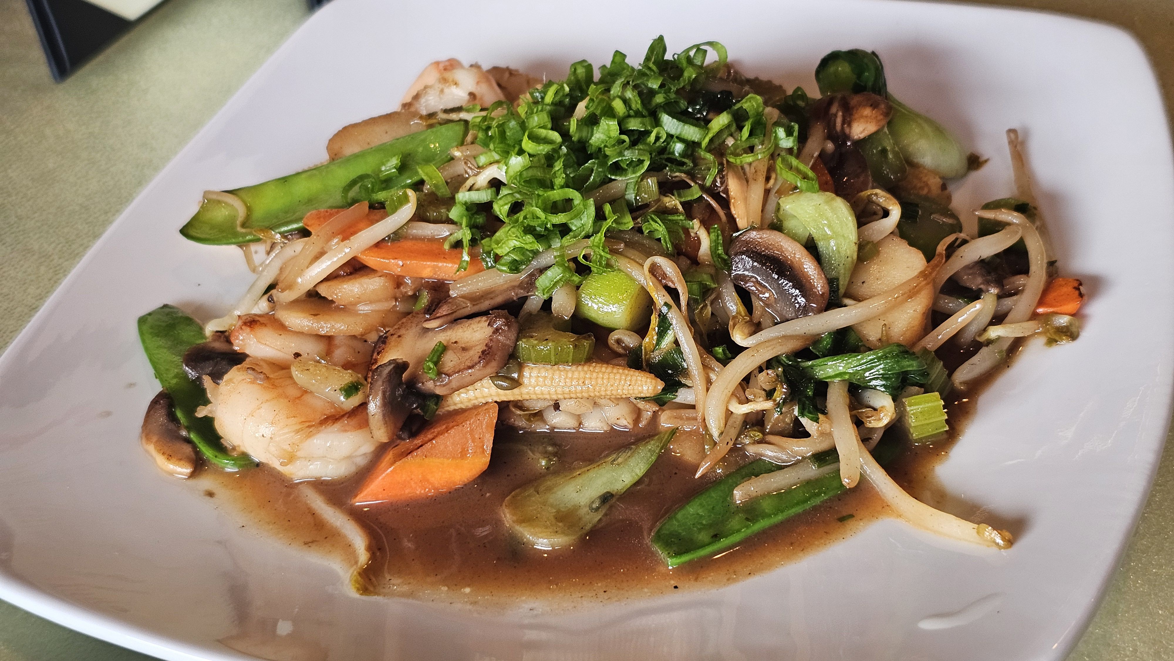 Stir-fried noodle dish with shrimp, mushrooms, carrots, snap peas, bok choy, bean sprouts, and chopped green onions in a brown sauce on a white plate.