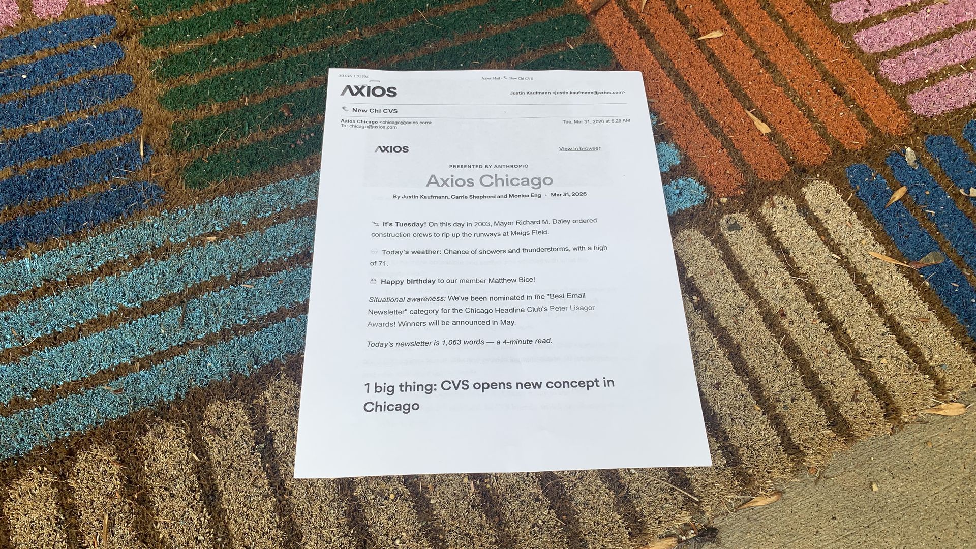 White Axios Chicago newsletter sheet on a colorful outdoor doormat with blue, green, orange, and pink stripes; sunlit with a few dry leaves around.