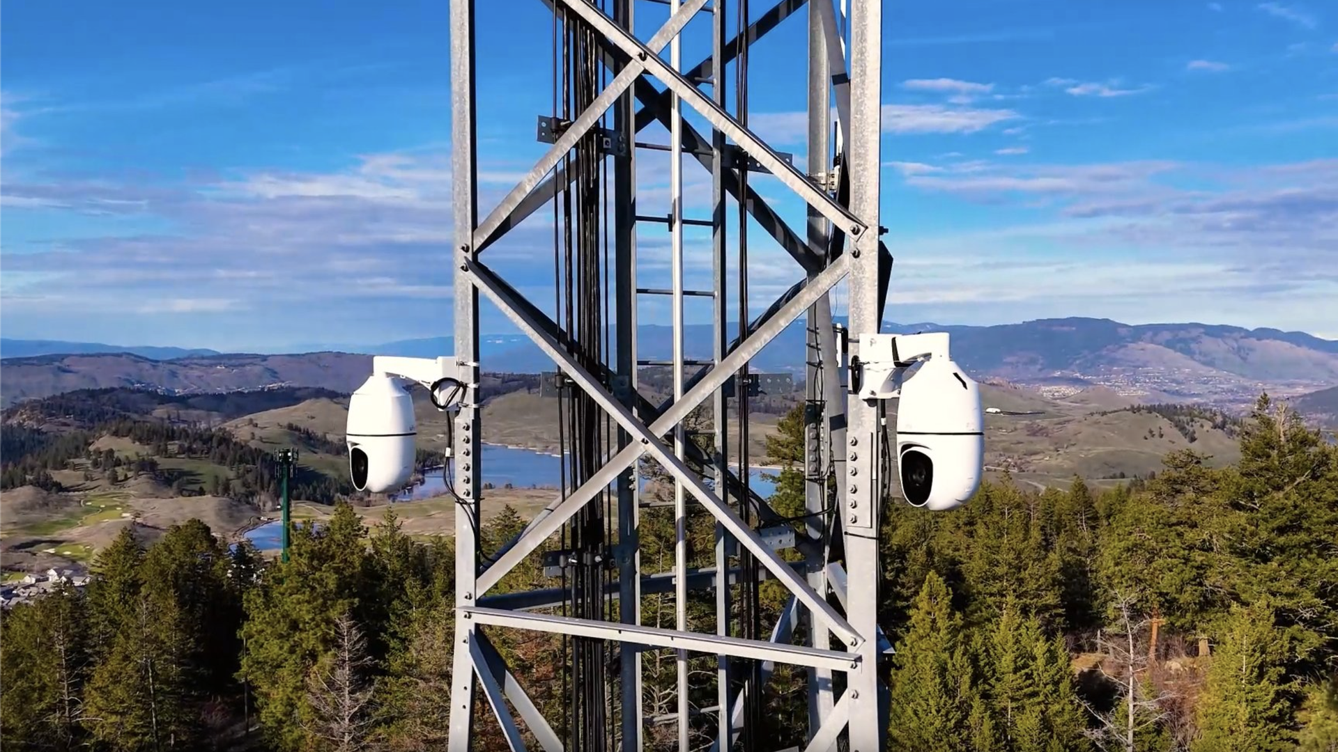 Close-up of a metal tower with two white security cameras mounted on either side, overlooking green trees, hills, a river, and mountains under a blue sky with scattered clouds.