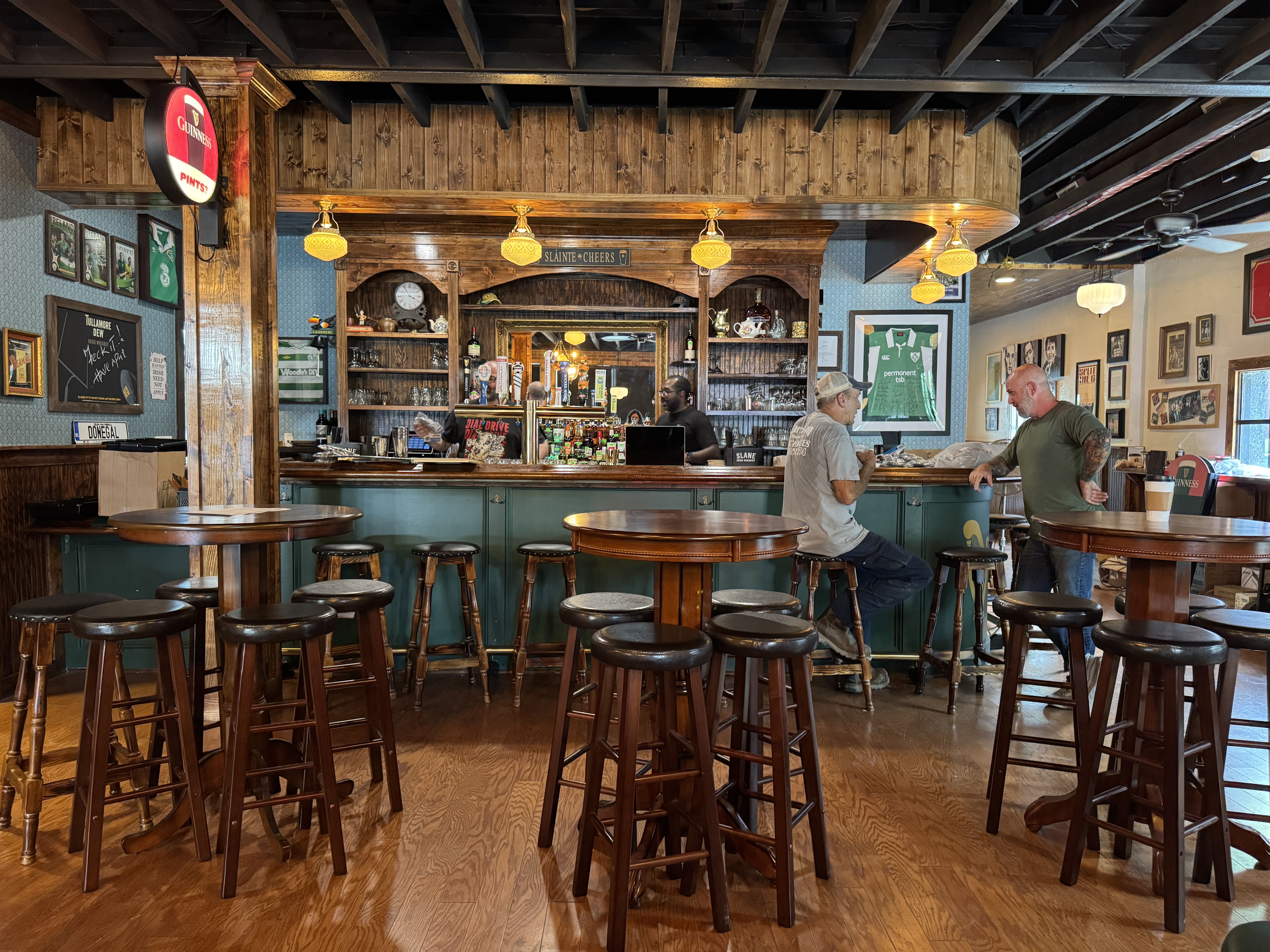 Interior of a cozy pub with wooden tables and stools, a green bar counter, two men talking near the bar, warm yellow lights, and framed sports memorabilia on blue walls.